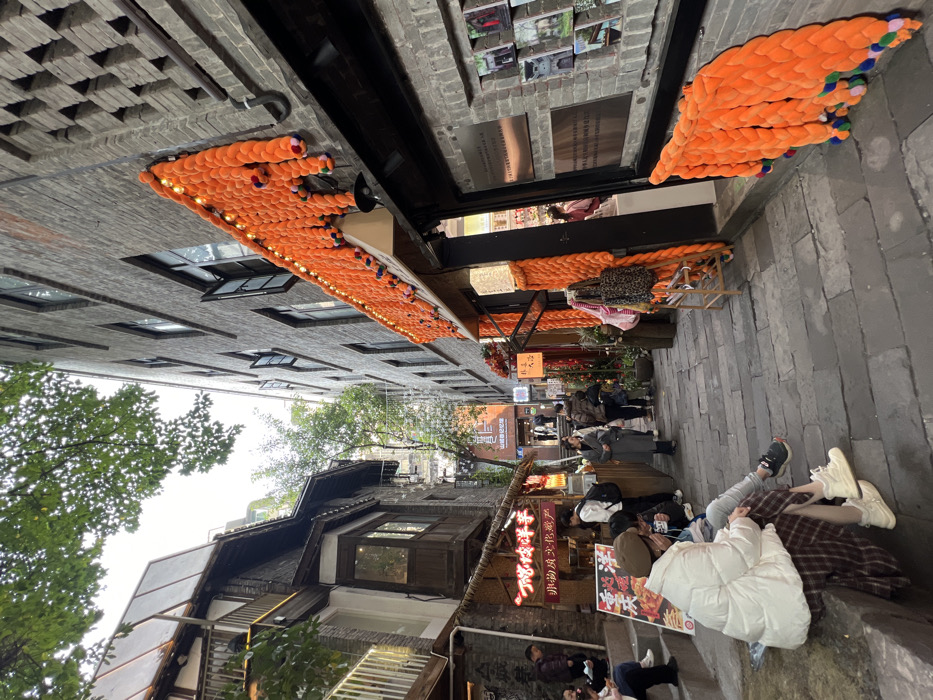 A narrow urban street scene with pedestrians. Traditional Chinese buildings line the sides, adorned with illuminated signage. Overhead, orange yarn art hangs between buildings, creating a visually striking canopy. People sit on steps, with a white puffy jacket prominent in the foreground. The composition features a mix of architectural styles and elements of modern urban life in a historic setting.