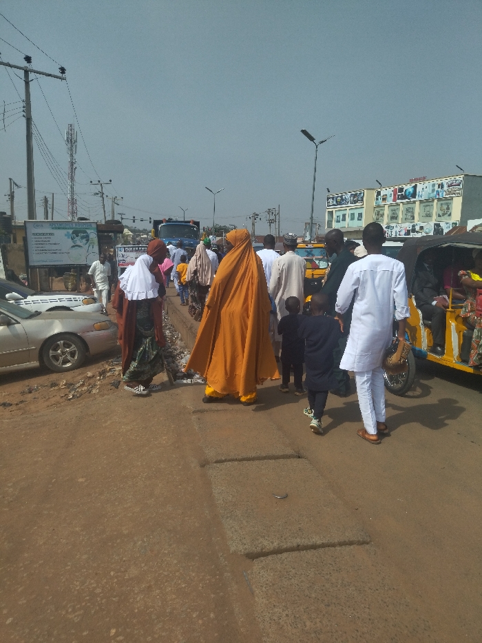 The scene is a street in New Karu, Nigeria, a suburban district in the Federal Capital Territory. It appears to be a busy street with a number of pedestrians walking in both directions, including several women dressed in traditional Muslim clothing. There is a large truck parked on the right side of the street and a yellow auto-rickshaw is driving towards the camera. The street is unpaved and there are several potholes. The background shows buildings with storefront signage. The weather appears clear and sunny. The time of day appears to be late afternoon. The location coordinates are latitude: 8.985, longitude: 7.565. A billboard advertises "Kwararafa Hospital" at the beginning of the street.