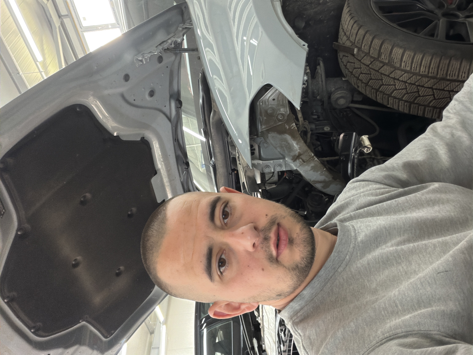 A close-up, angled shot depicts a man in a grey sweatshirt, positioned in front of an automobile with its hood open. The car's tire is visible in the upper right corner, while the engine components are partially visible in the background. The location appears to be a garage or workshop, identifiable by the overhead lighting fixtures. The city and specific location are unknown.