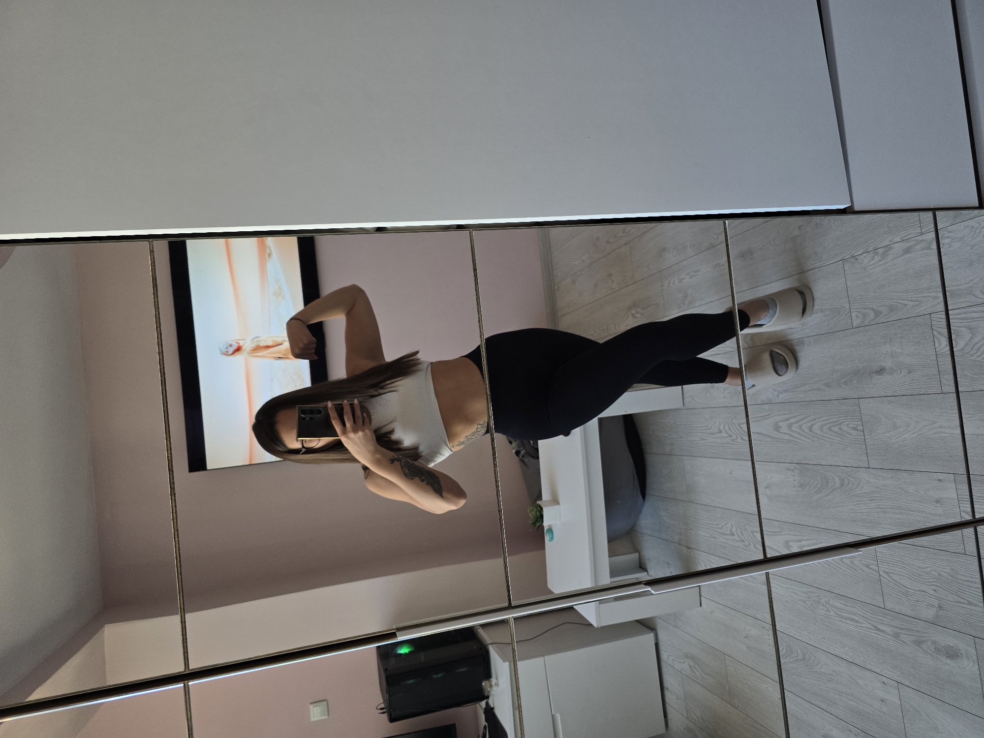 A young woman is posing for a photograph in a room with pink walls and light-colored wood flooring. She is wearing a white top and black leggings, with light-colored socks or slippers on her feet. She is holding a smartphone in front of her face to take a selfie, flexing her right arm. A television is visible on the wall behind her, with a light-colored image displayed. A white cabinet or shelf unit is positioned next to her. The image is a reflection in a mirrored surface, which has vertical seams, suggesting a wardrobe or a series of mirrored panels. An air conditioning unit and an electrical outlet are visible low on the wall. The city and country are unknown and cannot be inferred from the image.