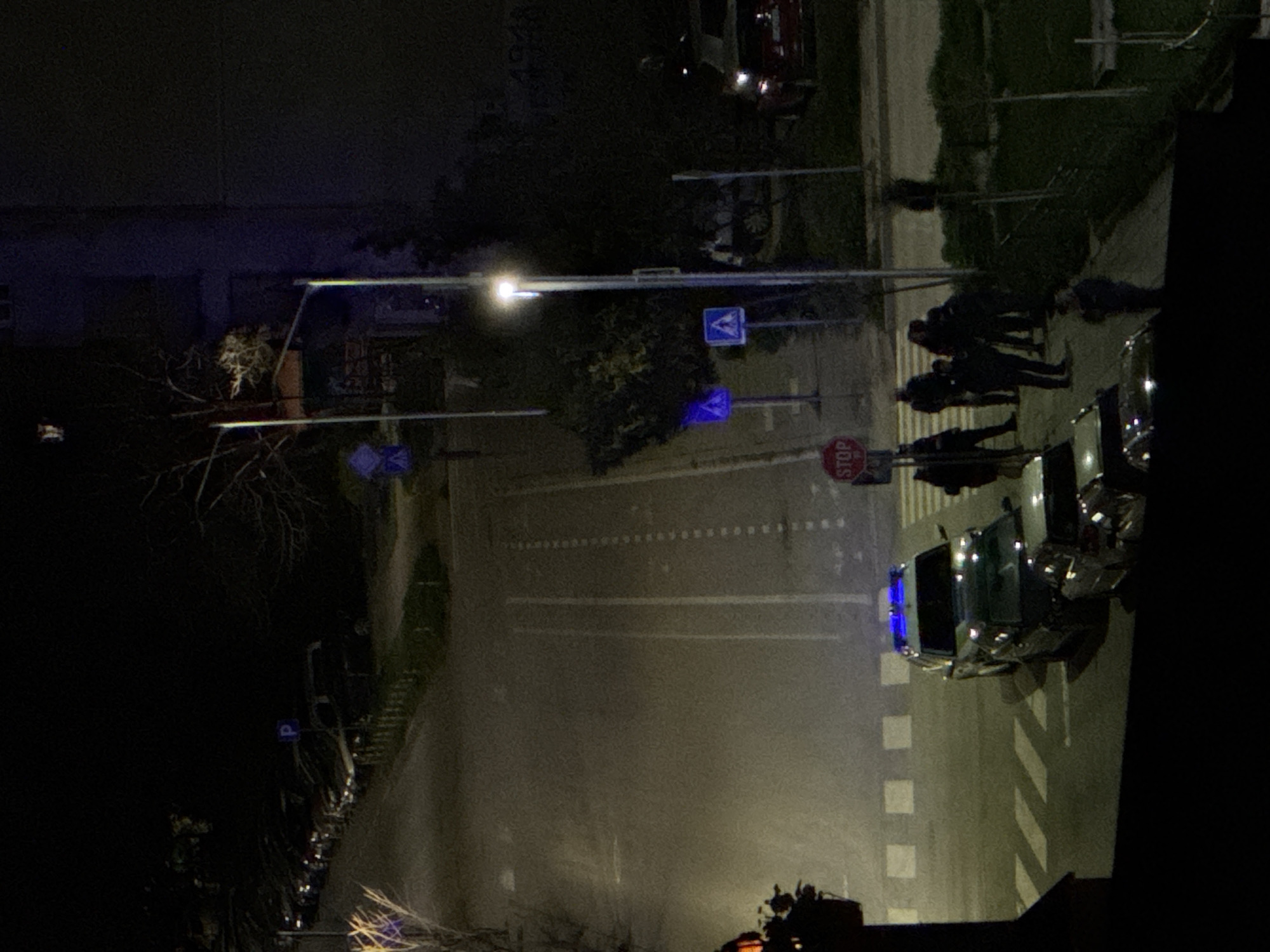 The image depicts a street scene at night in Varna, Bulgaria. A group of people stands near a crosswalk next to a row of parked cars and a police vehicle. Streetlights illuminate a road with white lane markings and a stop sign. Traffic signs are visible along the street. Buildings and trees are present in the background.