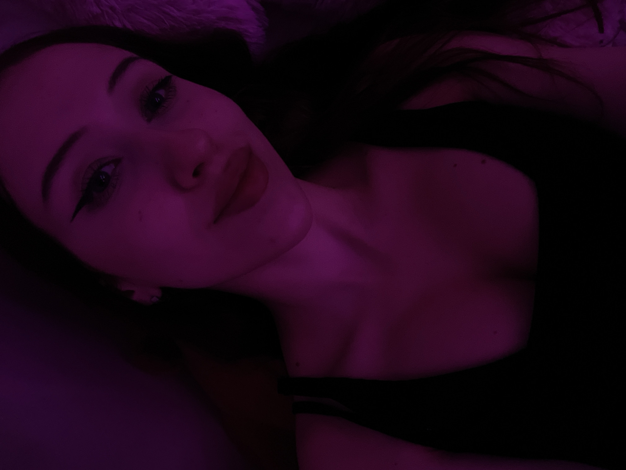 A young woman is shown in a close-up, overhead shot, illuminated by purple light. She is lying down and looking towards the camera, with her head slightly tilted. Her makeup is visible, including eyeliner and mascara. Her lips are slightly parted. She is wearing a black tank top. The background is dark, with a hint of a furry texture on the right side. There is no discernible urban or natural environment visible, and therefore no location within Votkinsk, Russia can be determined.