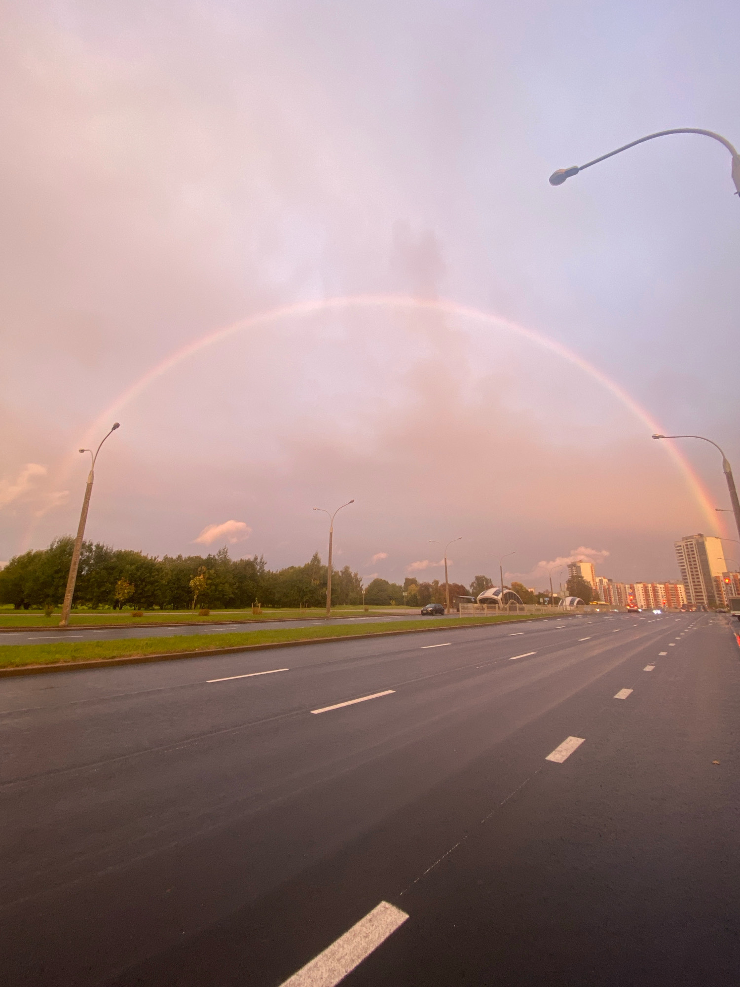 The image captures a wide asphalt road with white lane markings, likely a highway or a main city street, in Minsk, Belarus. The sky is overcast with a subtle pinkish hue, suggesting either dawn or dusk, and a faint rainbow arcs across the upper portion of the frame. Along the side of the road, there is a grassy median strip with trees and streetlights. In the distance, several modern buildings, including high-rise apartments, are visible. A few cars are present on the road, indicating moderate traffic. The overall scene depicts an urban landscape under atmospheric conditions, with the natural phenomenon of a rainbow adding a striking visual element.