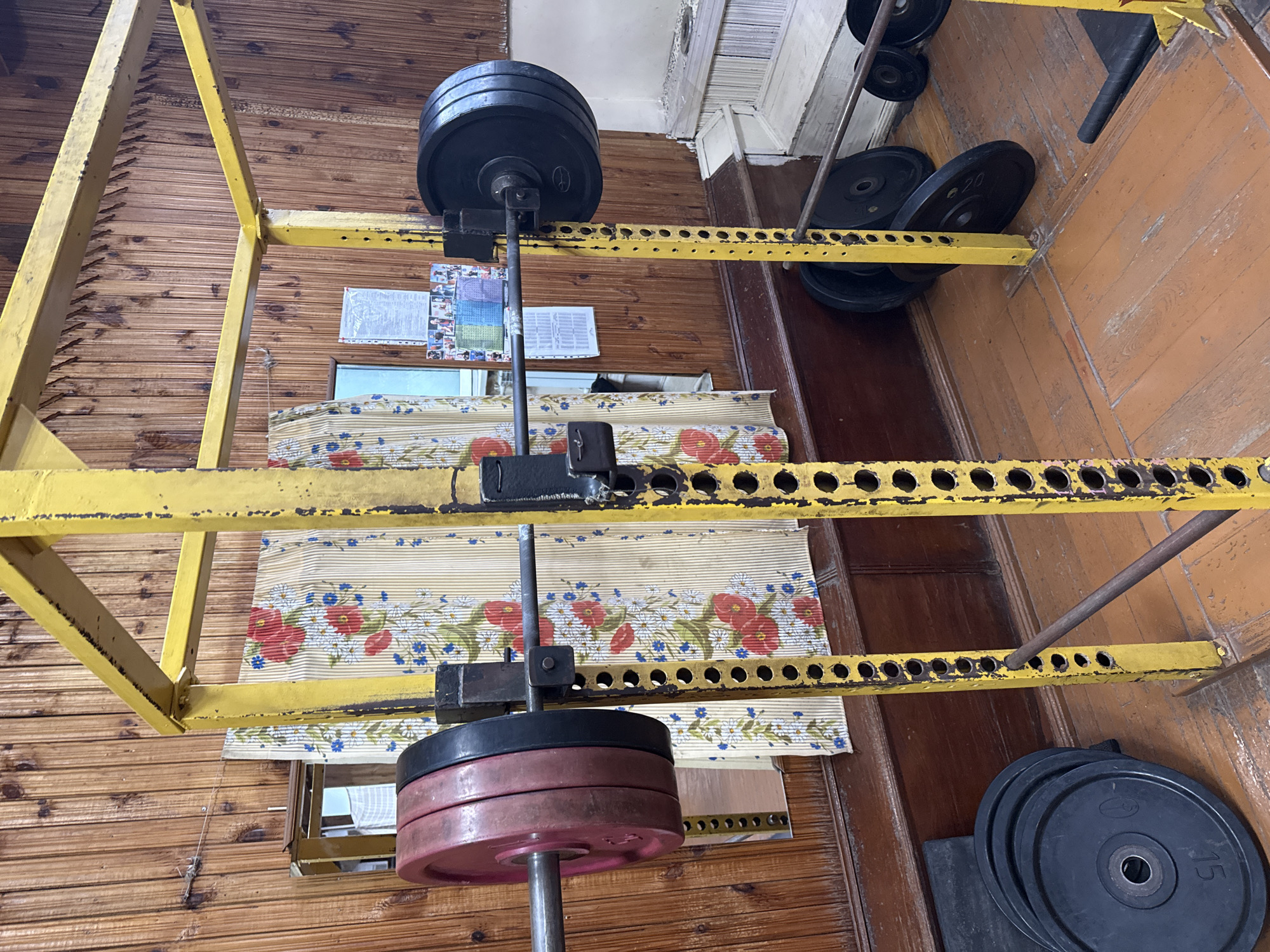 This image depicts a squat rack with a barbell loaded with weights. The rack is yellow and shows signs of wear. The barbell has red and black weight plates on either side. In the background, there is a wooden paneled wall, a window with floral curtains, and some papers pinned to the wall. The floor is made of wooden planks. There are additional weight plates on the floor to the left and right of the squat rack. This appears to be an indoor home gym setting, possibly in an older building. There are no people visible in the image. The location is not specified to be in Kazan, Russia.