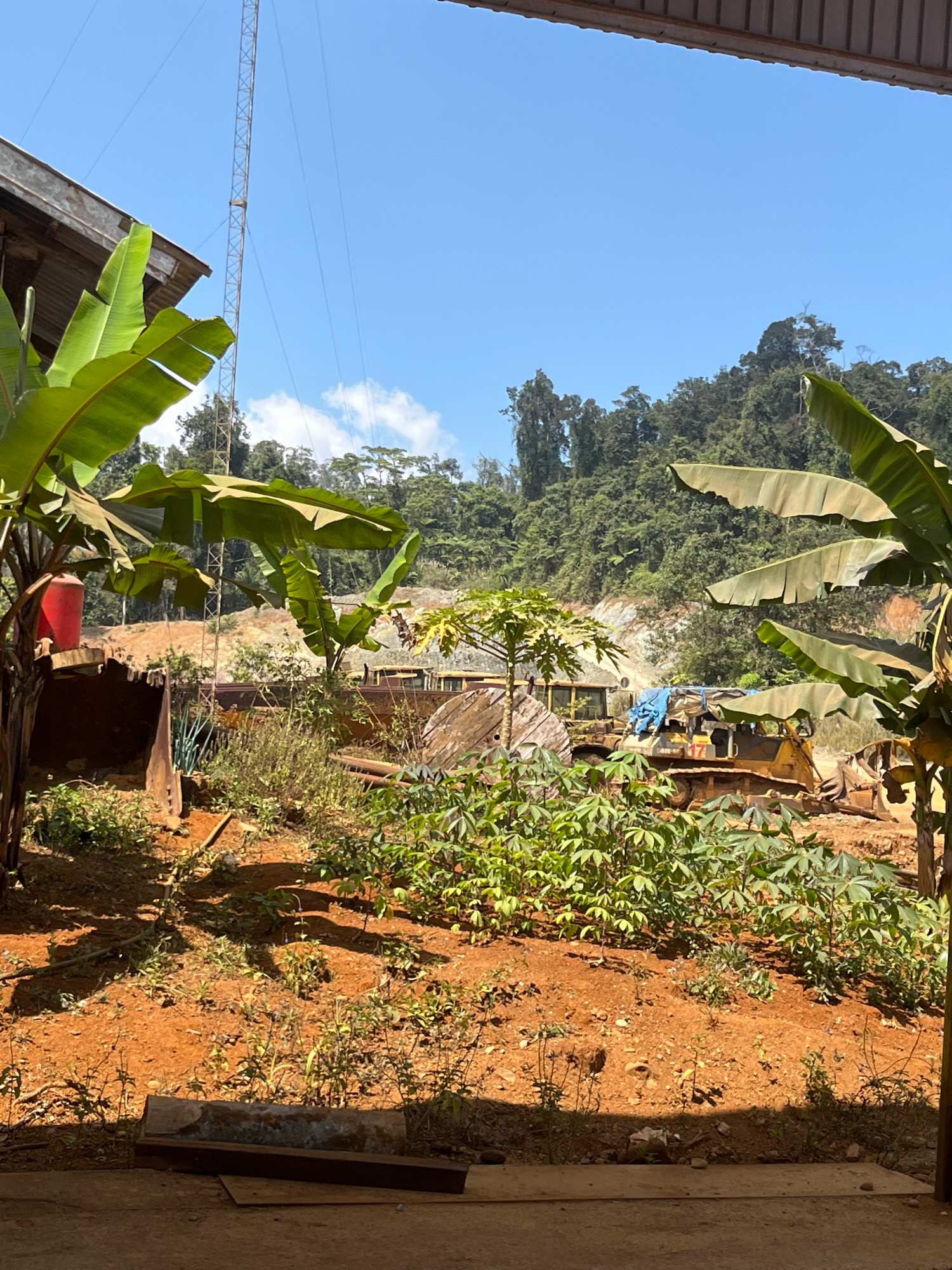 The image depicts an outdoor scene in what appears to be a rural or industrial area with lush vegetation and a clear blue sky. In the foreground, there is reddish-brown soil with some sparse greenery and shadows cast by unseen objects. To the left, a large banana plant with broad leaves is visible, along with a red bucket on what looks like a piece of excavation equipment. Behind this, a tall metal communication tower rises into the sky. In the middle ground, there are several pieces of heavy machinery, including what appears to be a bulldozer covered with a blue tarp, and another yellow bulldozer to its right. There are also large wooden spools and other industrial debris scattered around. The background is dominated by a densely forested hillside, with evidence of recent excavation or construction work on its lower slopes. The overall impression is one of an operational or recently used work site in a tropical environment. The location is identified as Unknown, Indonesia.
