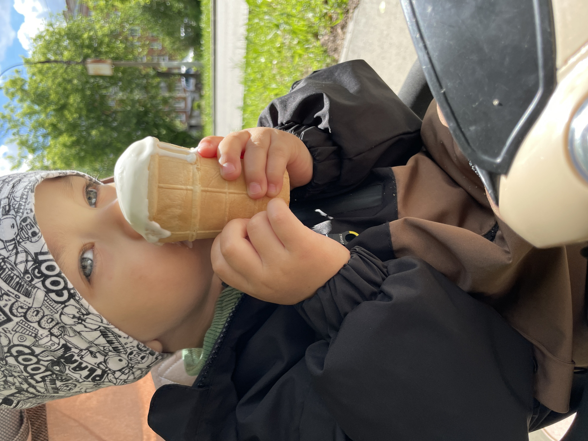 A young child is eating an ice cream cone while presumably seated in a stroller. The child has light skin, blue eyes, and is wearing a patterned head covering. The child is holding the ice cream cone with both hands. The child is wearing a jacket. Part of the stroller is visible on the right side of the frame. In the background are trees and a building, typical of an urban environment. There is no evidence to place this image in Izhevsk, Russia.