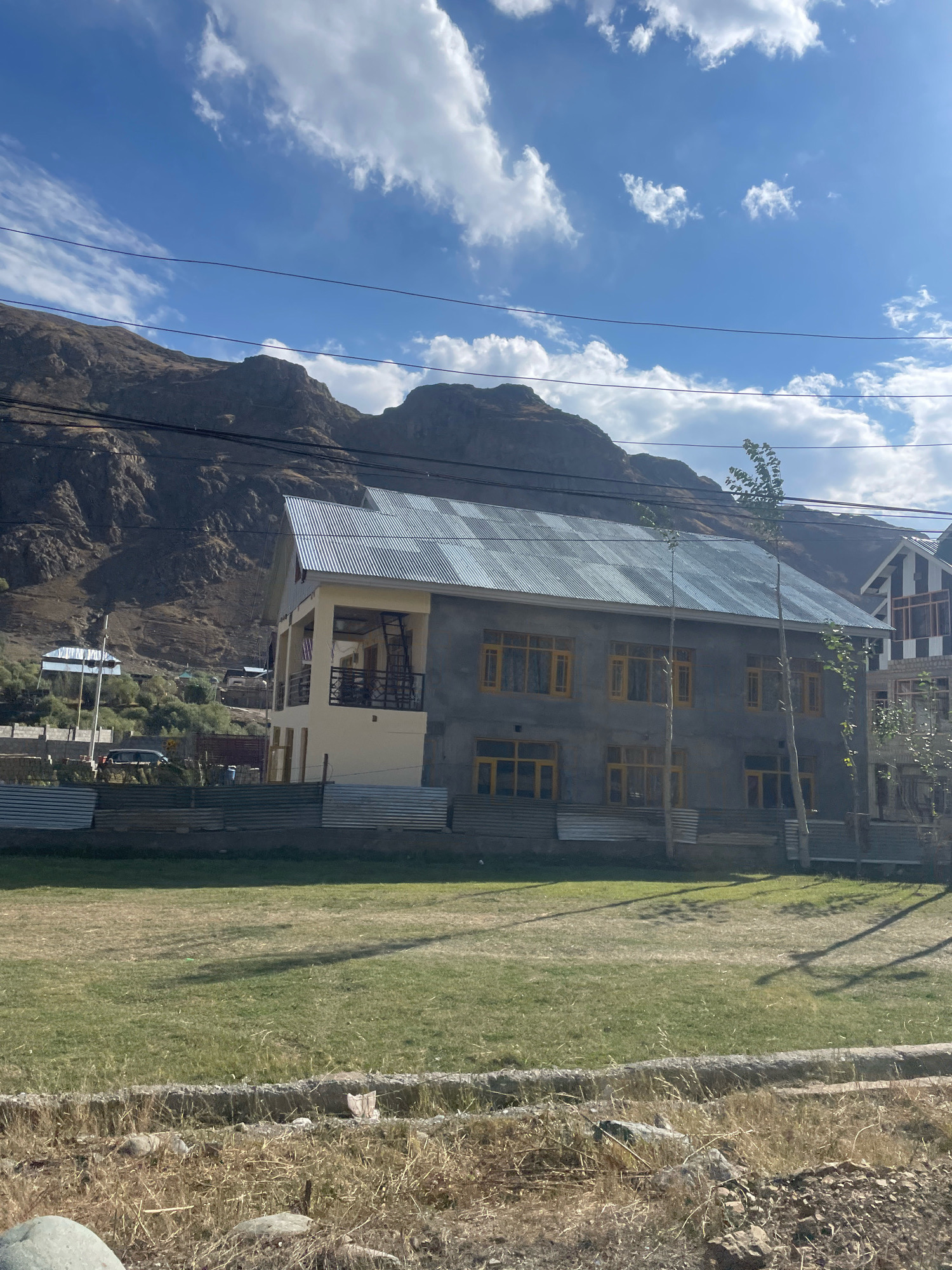 The image shows a building with a corrugated metal roof in Sokotyal, India. The building is two stories high and appears to be under construction or recently built, with unfinished grey concrete walls and yellow-framed windows. There is a balcony on the second floor. A large mountain range with sparse vegetation forms the backdrop, and the sky is blue with scattered white clouds. In the foreground, there is a grassy field and a fence made of corrugated metal sheets. A few small trees are planted in front of the building. The scene is illuminated by natural daylight.