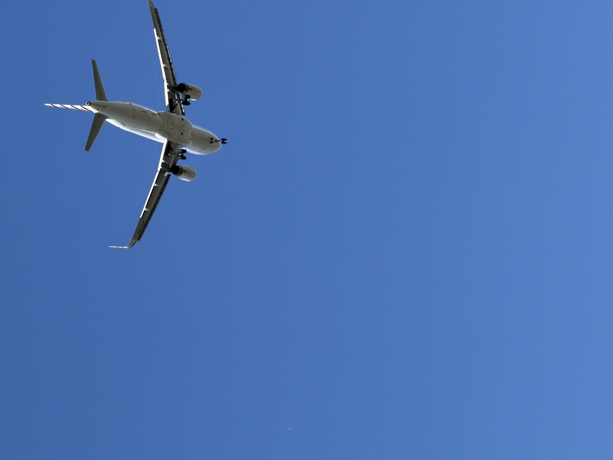 The media file displays a low-angle shot of a passenger airplane in flight against a clear, bright blue sky. The airplane is the primary object, positioned in the upper left quadrant of the frame. It is seen from below, revealing its underside, engines, landing gear extended, wings, and tail. The airplane has a white fuselage with red and white stripes on the tip of its tail fin. A faint, small, white speck, possibly a distant cloud or a lens flare artifact, is visible in the lower center of the sky. The overall scene suggests daylight, with the sun likely high in the sky given the strong illumination and the deep blue of the sky. The setting is an open sky, typical of an aerial view near an airport or during a flight. No people are visible. The activity depicted is the flight of the airplane. There is no visible text in the image. The location context provided is Burgas, Bulgaria.