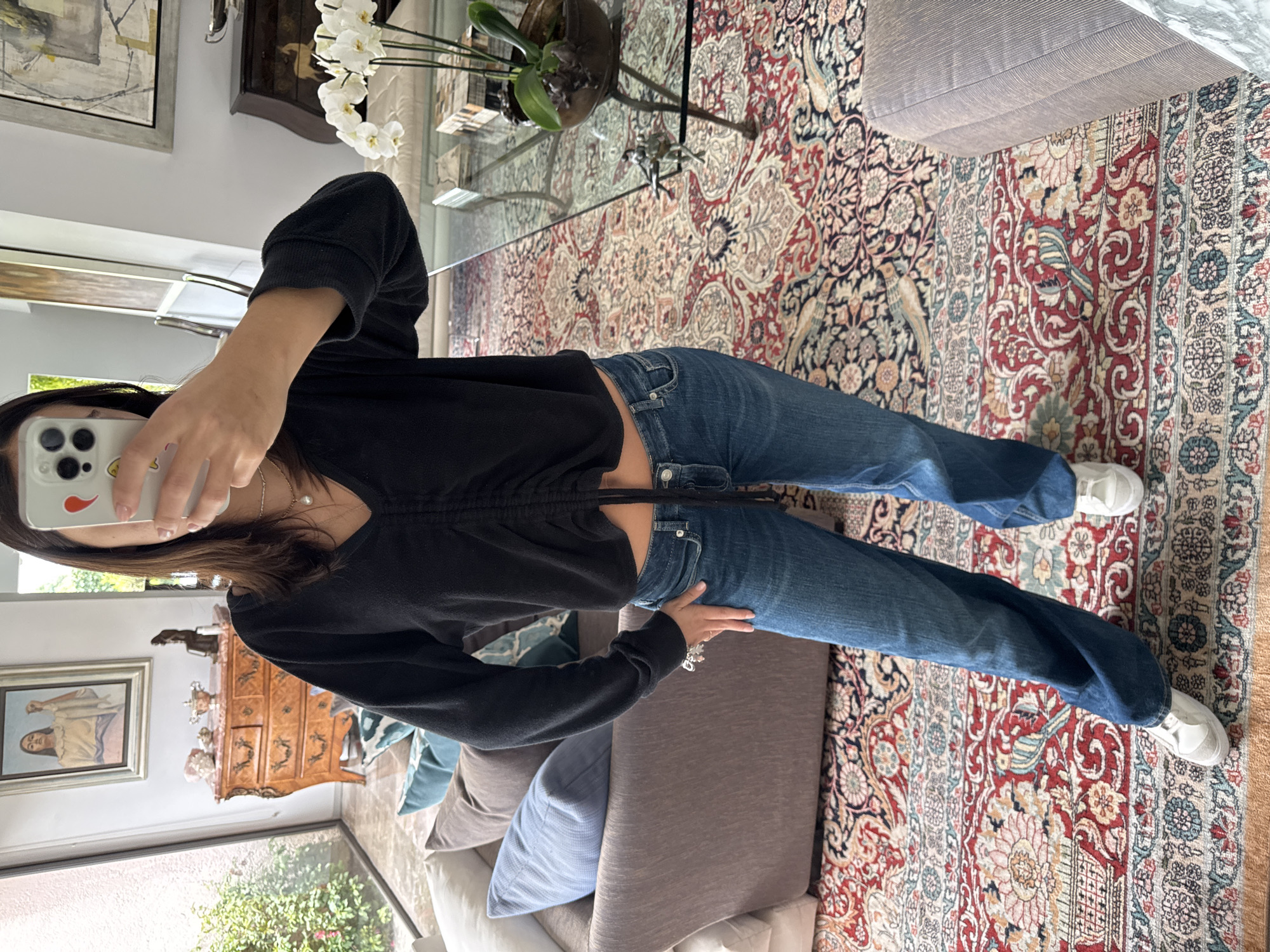 A woman is depicted taking a mirror selfie in what appears to be a living room. She is positioned slightly to the left, capturing her reflection from the chest down. She wears a black long-sleeved top with a v-neck and a central drawstring detail, blue jeans, and white sneakers. A thin gold necklace with a small pearl pendant is visible around her neck. Her face is mostly obscured by the white-cased iPhone she holds up, which features a yellow smiley face sticker and a red pepper sticker on the back.
The background reveals a richly decorated indoor space. A prominent feature is the large, ornate patterned rug covering the floor, displaying intricate floral, paisley, and possibly bird motifs in shades of red, cream, blue, and black. To the right, a glass coffee table holds a potted plant with white orchids. Behind it, an antique-style wooden chest of drawers with ornate handles and carvings is adorned with decorative vases and small floral arrangements. Above the chest, a framed painting of a woman is visible. On the left side of the room, another abstract framed artwork hangs on a light-colored wall. A sofa with various pillows in shades of gray and light blue is partially visible to the woman's right. The scene is illuminated by natural daylight, possibly from windows reflecting in the mirror. There is no visible text in the image.