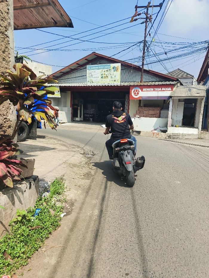 The image shows a man riding a black motorcycle on a paved street in Cileunyi, Indonesia. The motorcycle is moving away from the camera. The man is wearing a black shirt and blue jeans. He is wearing a helmet. The street is lined with trees and bushes. There is a building on the left side of the image. There is a sign on the building that reads "TR. BIRMAH BIRI JAYA." The building is also adorned with red brick, likely decorative. The road appears to be a narrow side street, with a curb and a sidewalk, likely a residential area. The location is at the intersection of two roads, a small junction. The image is taken from a low angle, showing the man's back and the motorcycle from the rear. The sky is blue and clear. There is a large utility pole and many power lines running overhead.
