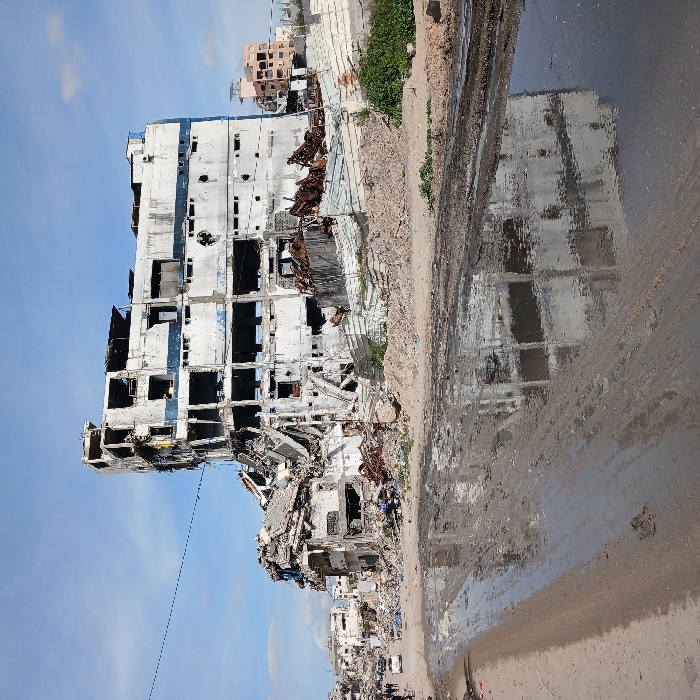 The image depicts a scene of extensive structural destruction in Jabalia, Palestinian Territory, under a partially cloudy blue sky. In the center, a severely damaged multi-story concrete building stands, its facade largely blown out, revealing internal floors and exposed rebar. The building, originally light-colored with blue accents, is heavily soiled and fragmented, with large sections of its walls and roof missing or collapsed. To the left, another collapsed structure is visible, surrounded by immense piles of concrete rubble, twisted metal, and general debris extending into the background, where more damaged buildings are discernible. In the far right background, a less damaged multi-story building with an orange-brown facade is partially visible. The foreground features an unpaved, muddy road with large puddles reflecting the damaged structures and the sky. On the left side of the road, in the midground, several dark figures, likely individuals, are observed near a white sedan. Sparse green vegetation grows amidst the rubble on the right. A thin, dark line, possibly a utility cable, stretches across the upper portion of the frame.