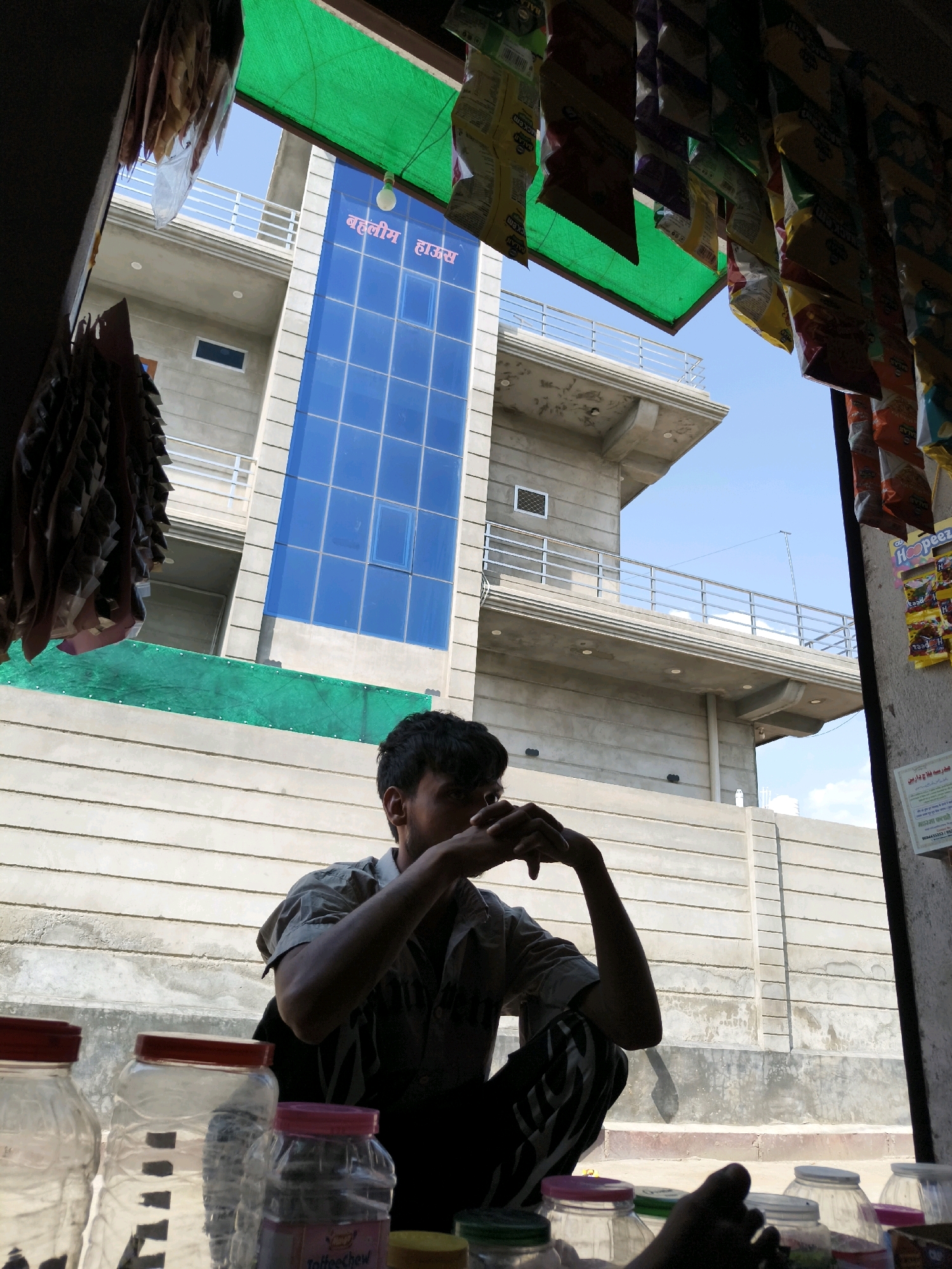 In Taranagar, India, a young man with dark hair sits on a low concrete ledge, his arms crossed and hands clasped in front of his face, suggesting contemplation or weariness. He is wearing a light-colored, short-sleeved shirt and dark pants with white stripes. In the foreground, several transparent plastic jars with colored lids are visible. Behind the man, a modern building with a striking blue glass facade rises against a clear blue sky. The building's name, "बहालीम हाउस" (Bahalim House), is prominently displayed in red lettering on the blue glass. Green tarpaulin is stretched across the top of the scene, partially obscuring hanging packets of merchandise, suggesting this is the entrance to a shop or stall. The overall environment appears to be an urban or semi-urban setting, with a mix of older, textured concrete walls and newer construction.