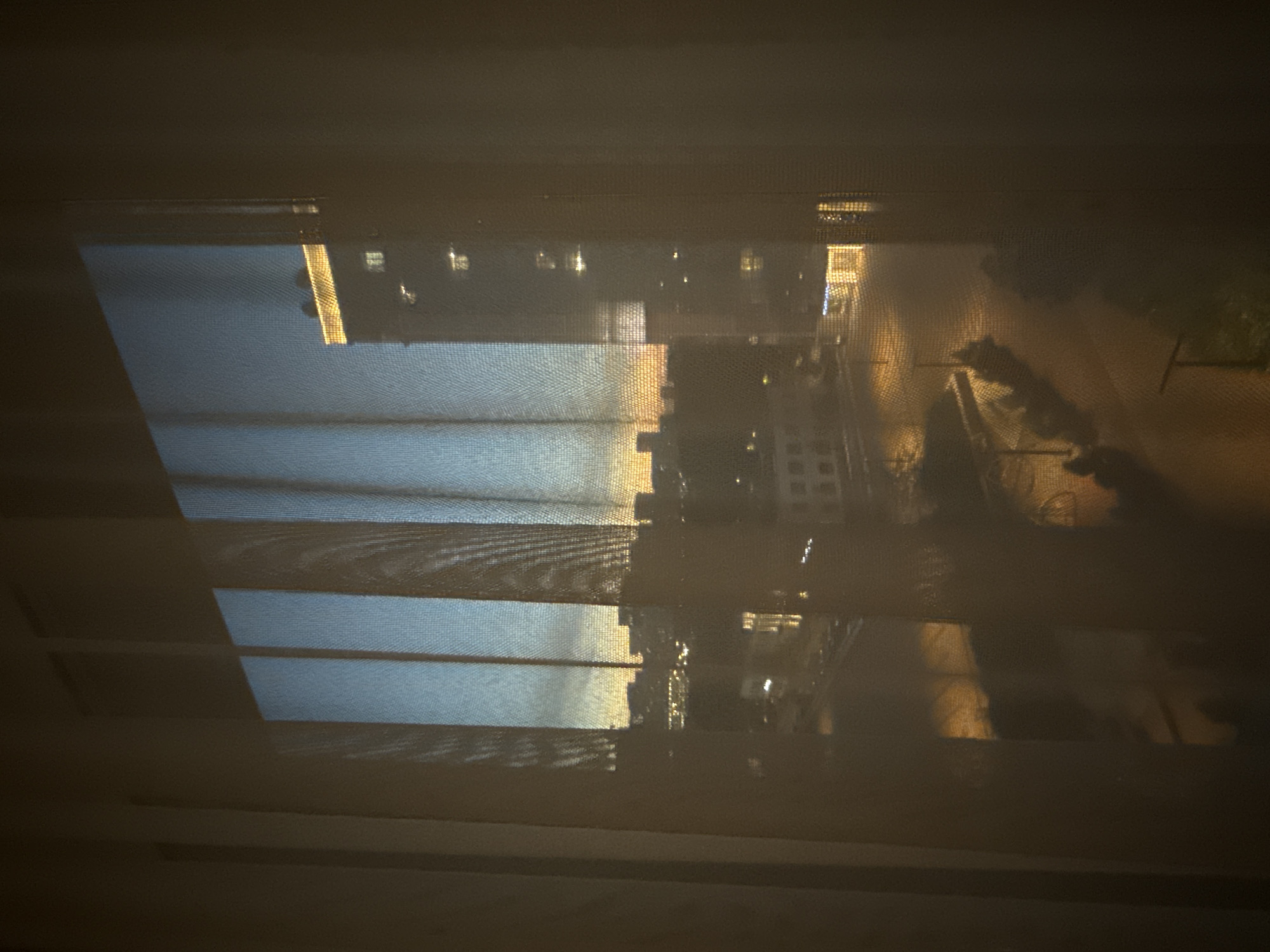 The image captures a view of a city at dusk, seen through sheer curtains. The sky exhibits a gradient of colors, transitioning from a deep blue at the top to warm hues of orange and yellow near the horizon, indicative of sunset or twilight. In the foreground, vertical folds of the curtains create a textured overlay, partially obscuring the scene beyond.
The cityscape features a mix of buildings, some appearing as lower structures with illuminated windows, suggesting residential or commercial spaces. In the middle ground, a distinct taller building stands out, illuminated by warm yellow lights along its upper edge and scattered lights within its structure. This building's architecture suggests it could be a hotel or an office building.
The ground level of the cityscape reveals illuminated streets or pathways, with streetlights casting a yellowish glow on the pavement. A road or pathway appears to wind through the scene, bordered by illuminated decorative lighting or street lamps. Some shadows are cast on the ground, suggesting the presence of trees or landscaping elements.
The overall impression is one of an urban environment at the transition from day to night, with artificial lights beginning to dominate the scene as natural light fades. The perspective is from an elevated vantage point, looking out over the city. The context provided indicates this scene is located in the city of Khimki, Russia.