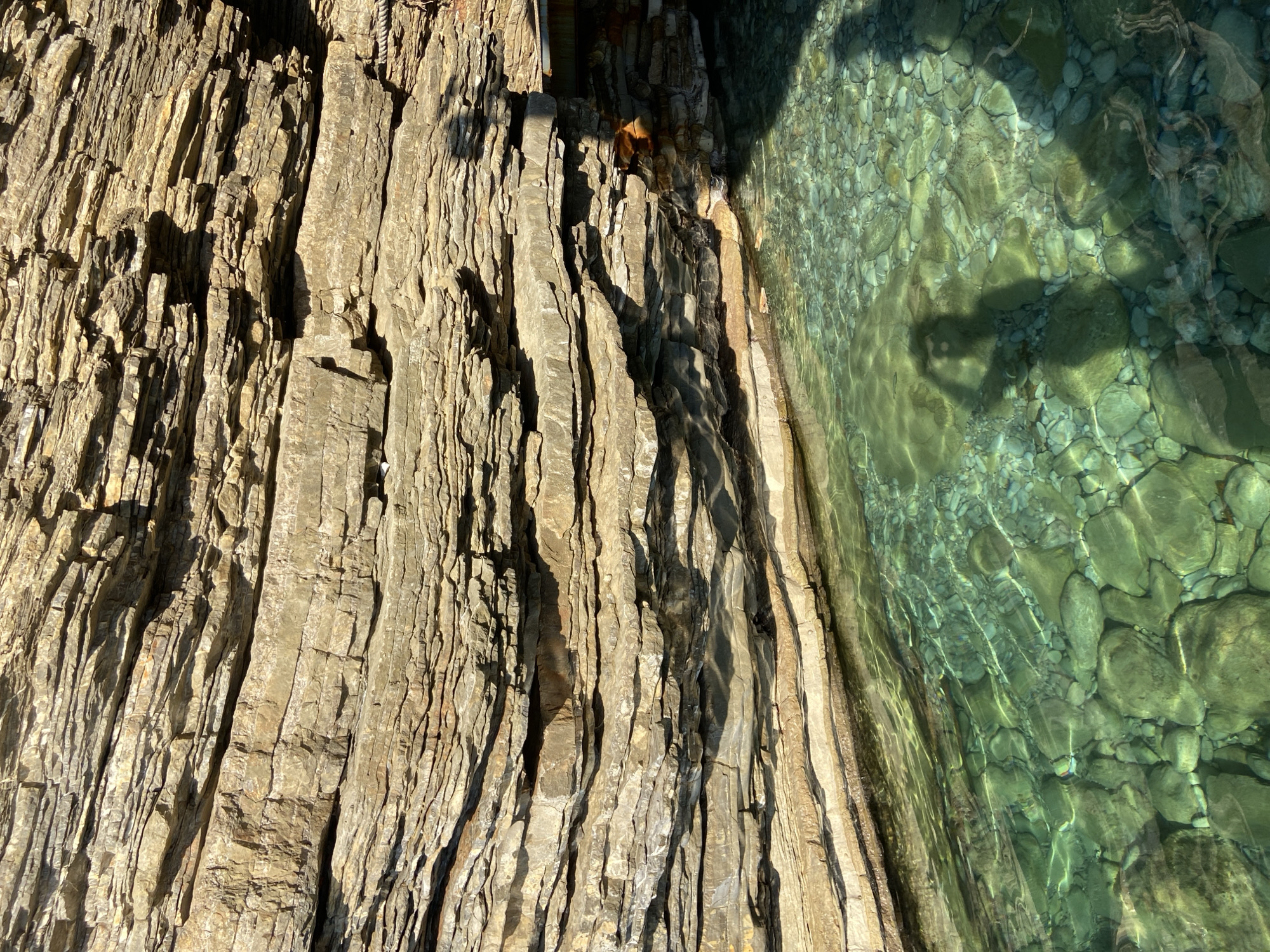 The image shows a natural rock formation adjacent to clear, shallow water filled with pebbles. The rock face is layered with shades of tan and brown, indicating a sedimentary geological structure. The water is a vibrant turquoise, with sunlight reflecting off its surface and illuminating the rocks below. The shadow of a person holding a phone or camera is visible in the water, suggesting the image was taken by someone standing at the edge of the water or slightly within it. A rope is faintly visible attached to the rock face on the right side of the image. In the upper right corner, a small section of what appears to be man-made construction, possibly a wooden platform or structure, is present. This scene is located at the Mogren Fortress, a historical fortification situated near the town of Budva, Montenegro.