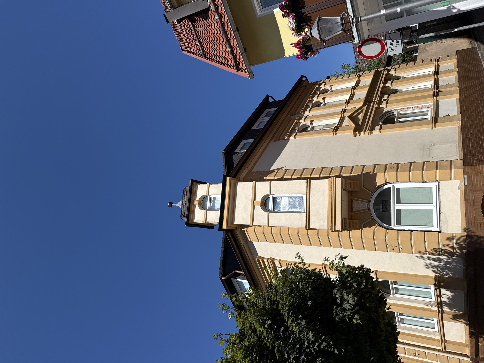 This is an outdoor, eye-level, medium shot photograph taken on a bright, sunny day in the city of Bad Sachsa, Germany. The photo features two buildings and a clear blue sky. The main building, on the left, is a multi-story structure with cream-colored walls and ornate yellow trim around the windows and corners. It has a dark roof and a small tower-like structure at the top with a weather vane. A large, leafy green tree is on the left side of the frame, partially obscuring the building. The second building, on the right, has a yellow facade and a red tiled roof. It features flower boxes with colorful flowers and a traditional-looking lamppost. A round traffic sign with a red border and a white center, indicating no entry, is visible on a pole in front of the yellow building, along with a rectangular sign below it that reads "Anlieger frei." The scene appears to be a street in a European town.