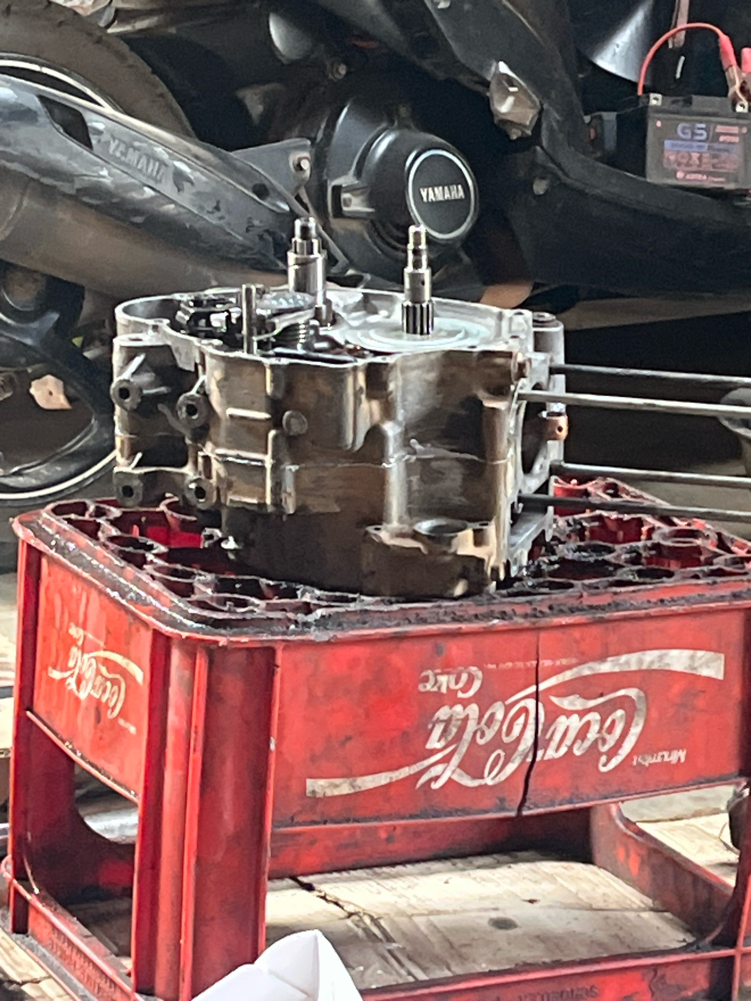 The image depicts a motorcycle engine case sitting on top of a red Coca-Cola crate. The engine case is metallic and appears to be disassembled, with various internal components visible. The Coca-Cola crate is made of plastic and has the Coca-Cola logo printed on its sides. In the background, parts of a motorcycle are visible, including a tire, the engine block, and a battery. The scene appears to be an outdoor workshop or garage, possibly in Kebumen, Indonesia, given the context of the request. The overall impression is one of a mechanical repair or maintenance activity.