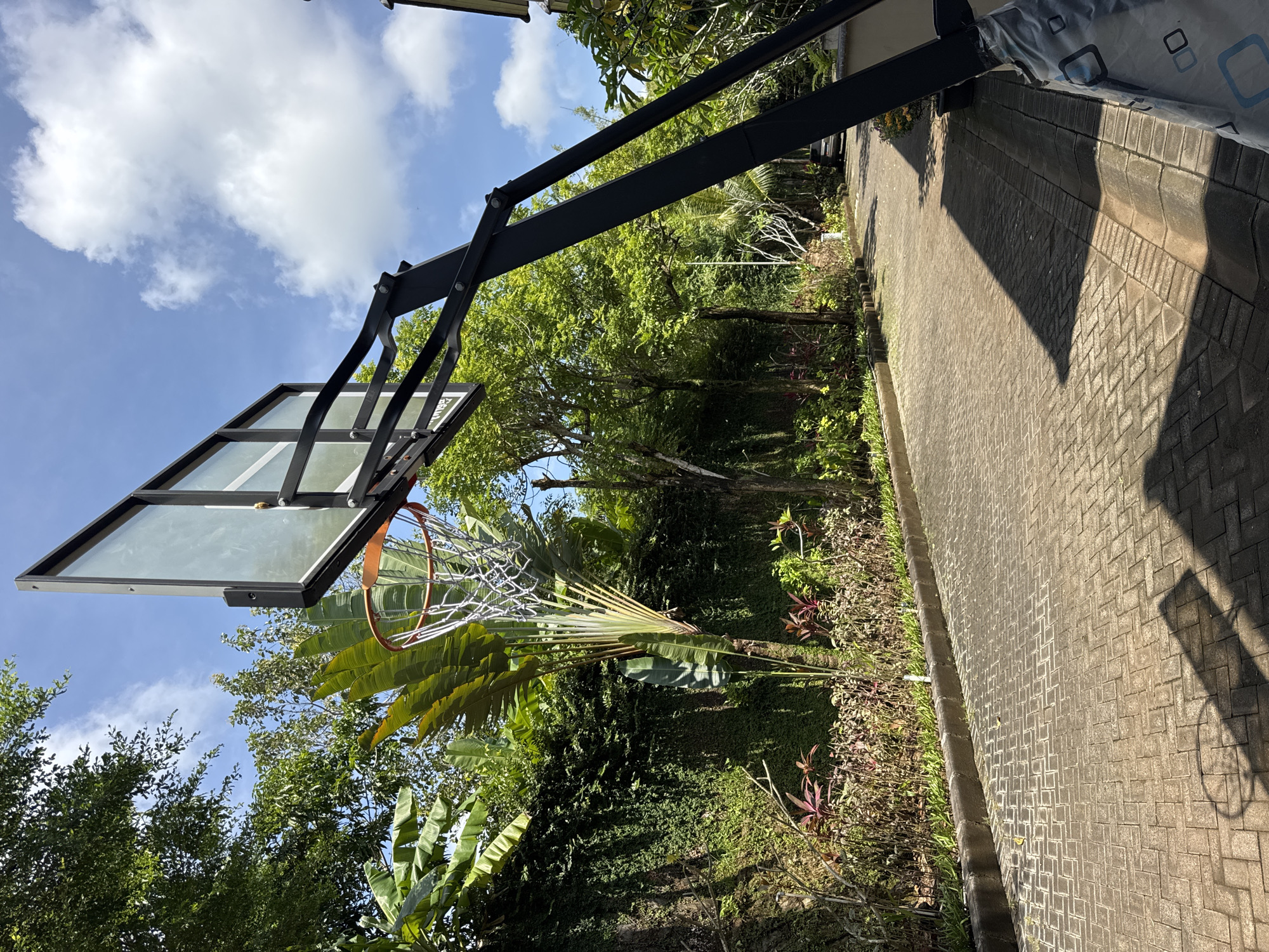 The image depicts a residential scene, likely in Jimbaran, Indonesia given the vegetation. A basketball hoop and backboard dominate the left side of the image, positioned near a paved driveway that extends into a lush garden area. The garden contains a variety of tropical plants, including palm trees and other greenery characteristic of the region. Above, a blue sky with scattered clouds is visible. The setting suggests a private residence with outdoor recreational space and landscaping typical of a tropical environment.