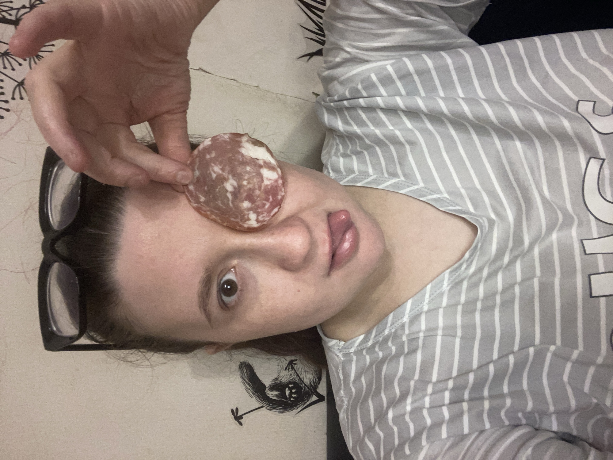 A person is lying down, holding a slice of salami over one eye. The person has dark hair and is wearing glasses. They are sticking their tongue out. The person is wearing a striped shirt. The background includes floral patterns and a drawing of a bird. The location is not discernible from the image. The city of Chaikovsky, Russia, is not discernible from the image.