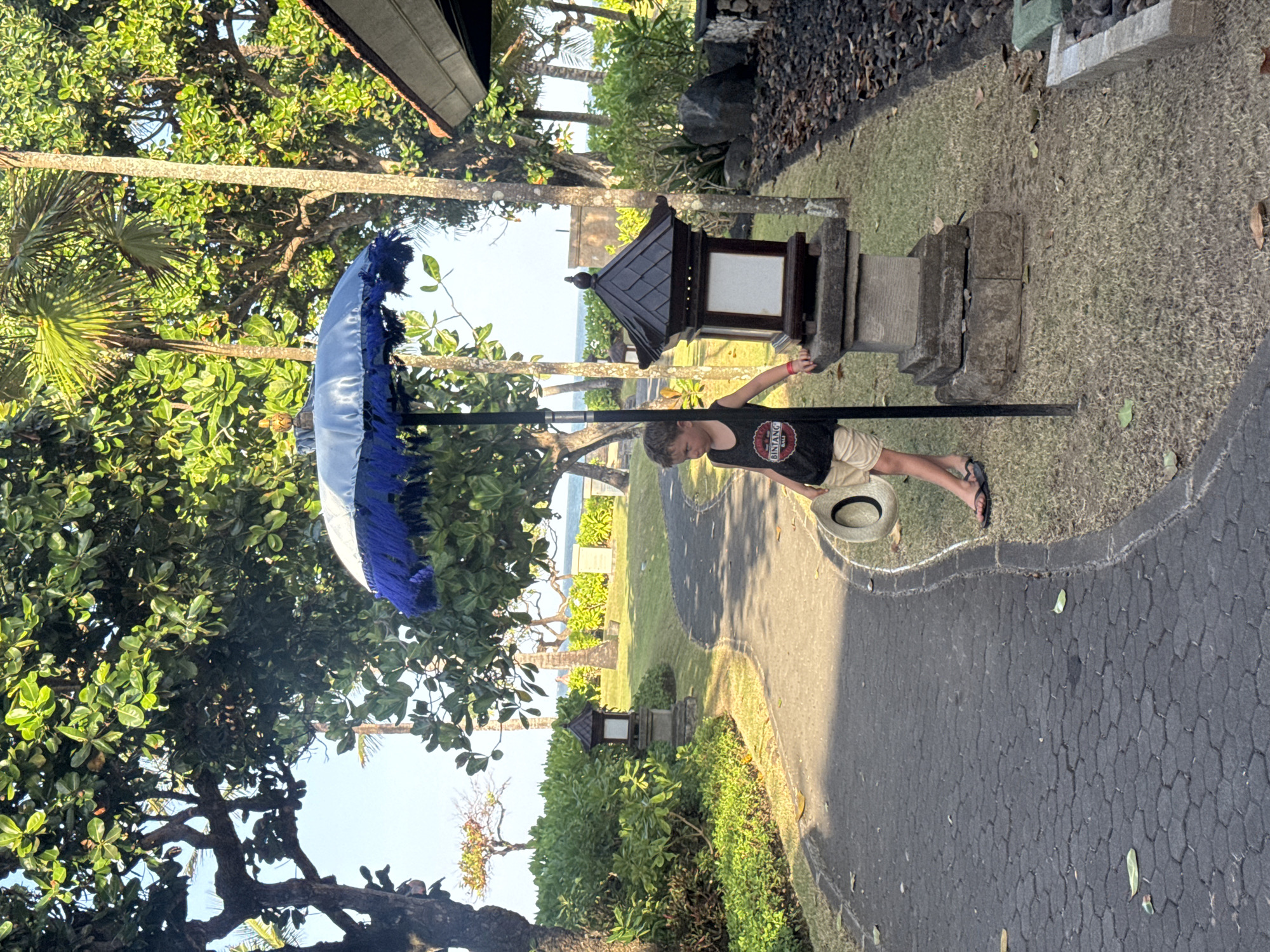 A young boy stands on a paved walkway in Kuta Selatan, Indonesia, holding a straw hat. He is wearing a black tank top with a circular red and white logo and light brown shorts. To his right, a dark brown, tiered lantern with a white panel sits on a stone pedestal. A blue and white umbrella with blue fringe is positioned behind the boy, supported by a black pole. The walkway curves to the left, bordered by green grass and lush vegetation. In the background, trees line a pathway leading towards the ocean, with the sea and sky visible in the distance. The scene is bathed in natural sunlight.