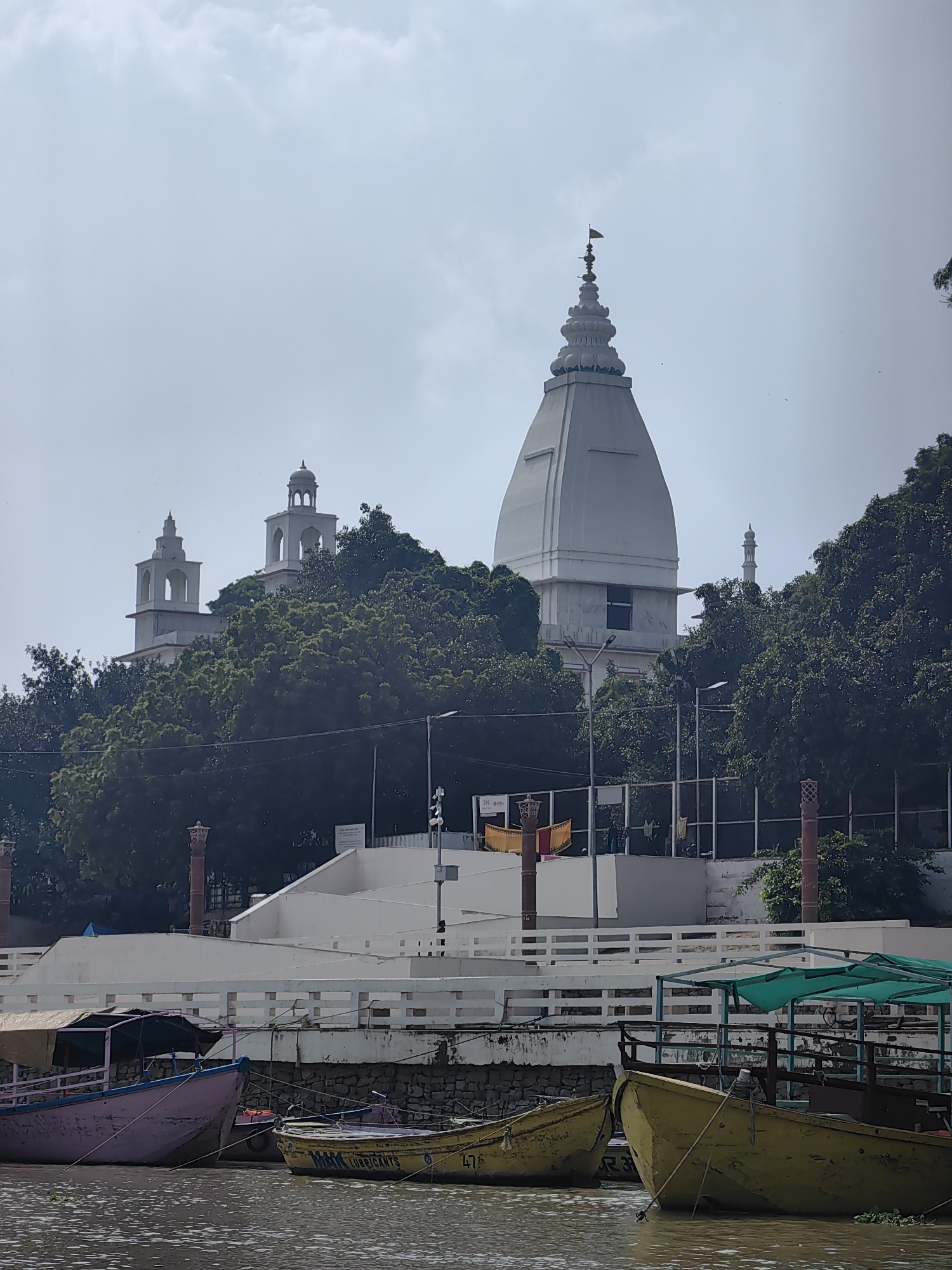 This image, captured on a cloudy day in Mughalsarai, India, depicts a riverside scene dominated by a large white temple structure. The temple's prominent feature is a conical spire topped with a small flag. To the left of the main spire, two smaller white towers with domed roofs rise above a dense canopy of green trees. In the foreground, several boats are moored on the murky water. One boat on the far left is painted pink and purple, with a faded tarp overhead. To its right is a yellow boat with "MAX LUBRICANTS" and the number "47" visible on its side. Another yellow boat, partly obscured, is tied to the pink boat. A green canopy stretches over a portion of a fourth boat on the right. A white, multi-tiered railing runs along the embankment behind the boats, with decorative pillars visible at intervals. Beyond the railing, a white building complex extends, and behind that, the temple complex is nestled amongst lush foliage. In the distance, a minaret-like structure is visible. The sky is overcast with light clouds, and a few birds can be seen in the sky. There are no visible people in the image.