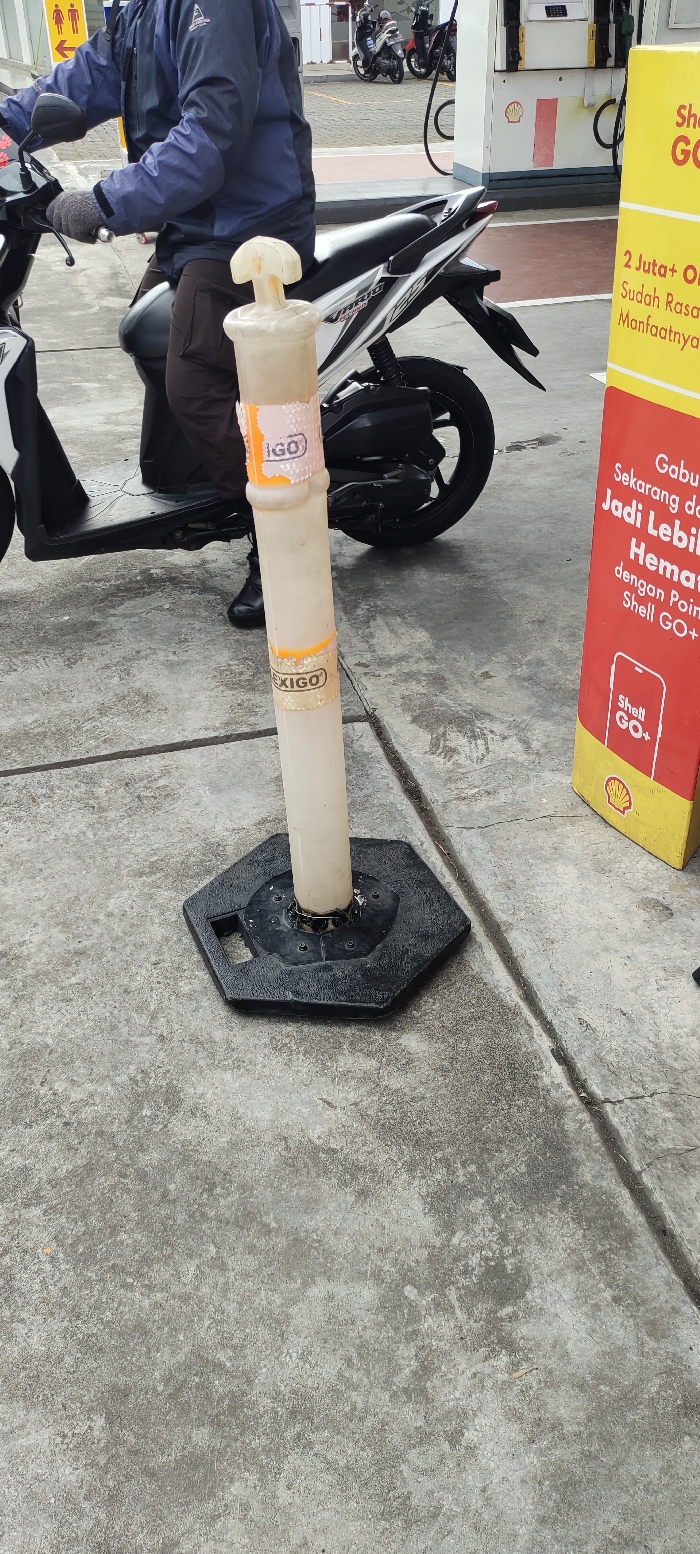 The image shows a close-up view of a white, cylindrical bollard with a black base located at a gas station in Kecamatan Lawang, Indonesia. The bollard is partially obscured by a person in a blue jacket riding a motorcycle, standing next to the bollard. The person's right leg is visible in the photo. The bollard is marked with two orange and black stickers that read "IGO". There is a black motorcycle in the background, with the front tire visible and a red and yellow sign on the right. The image is taken at a gas station with a concrete surface. The overall environment appears to be a gas station. The surface of the bollard is smooth. The bollard is made of a white plastic material. The bollard is a traffic control device. There is a slight difference in elevation between the pavement around the bollard and the pavement around the motorcycle. The motorcycle is parked. The motorcycle is facing the right of the image. The bollard is the main subject of the photo.