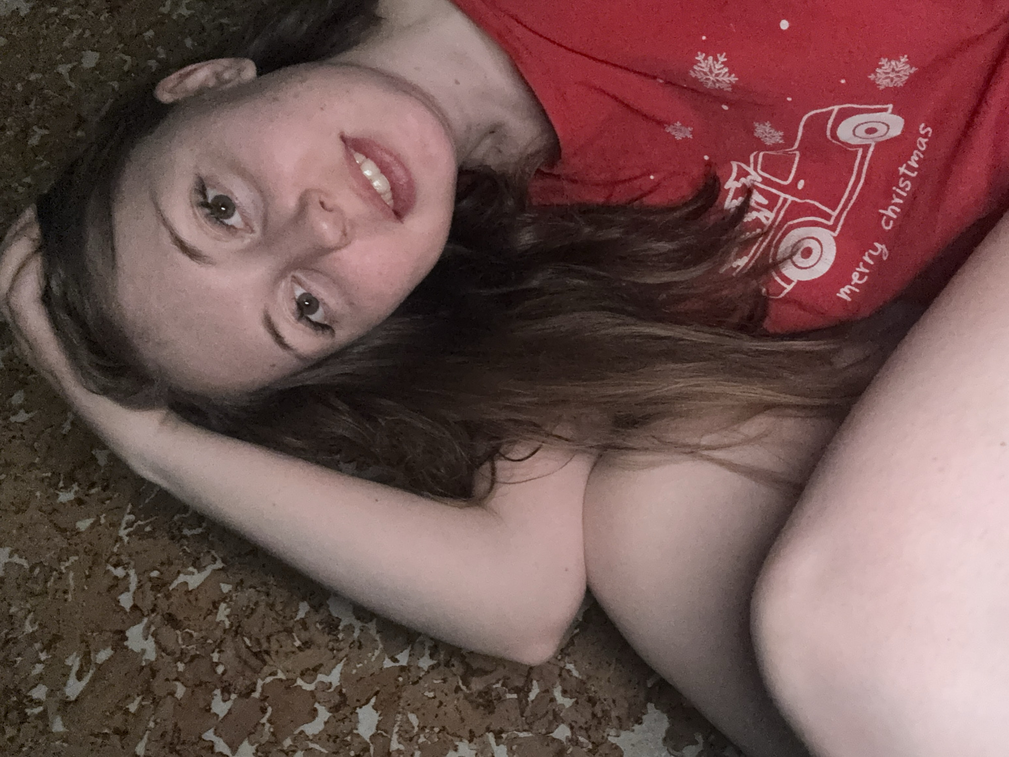 A young woman with brown hair is lying on a patterned surface. She is wearing a red t-shirt with a white graphic of a truck and the words "merry christmas". She is smiling slightly and looking at the camera. Her left arm is bent and her hand is near her head. Her right arm is extended near her body. The setting is indoors.