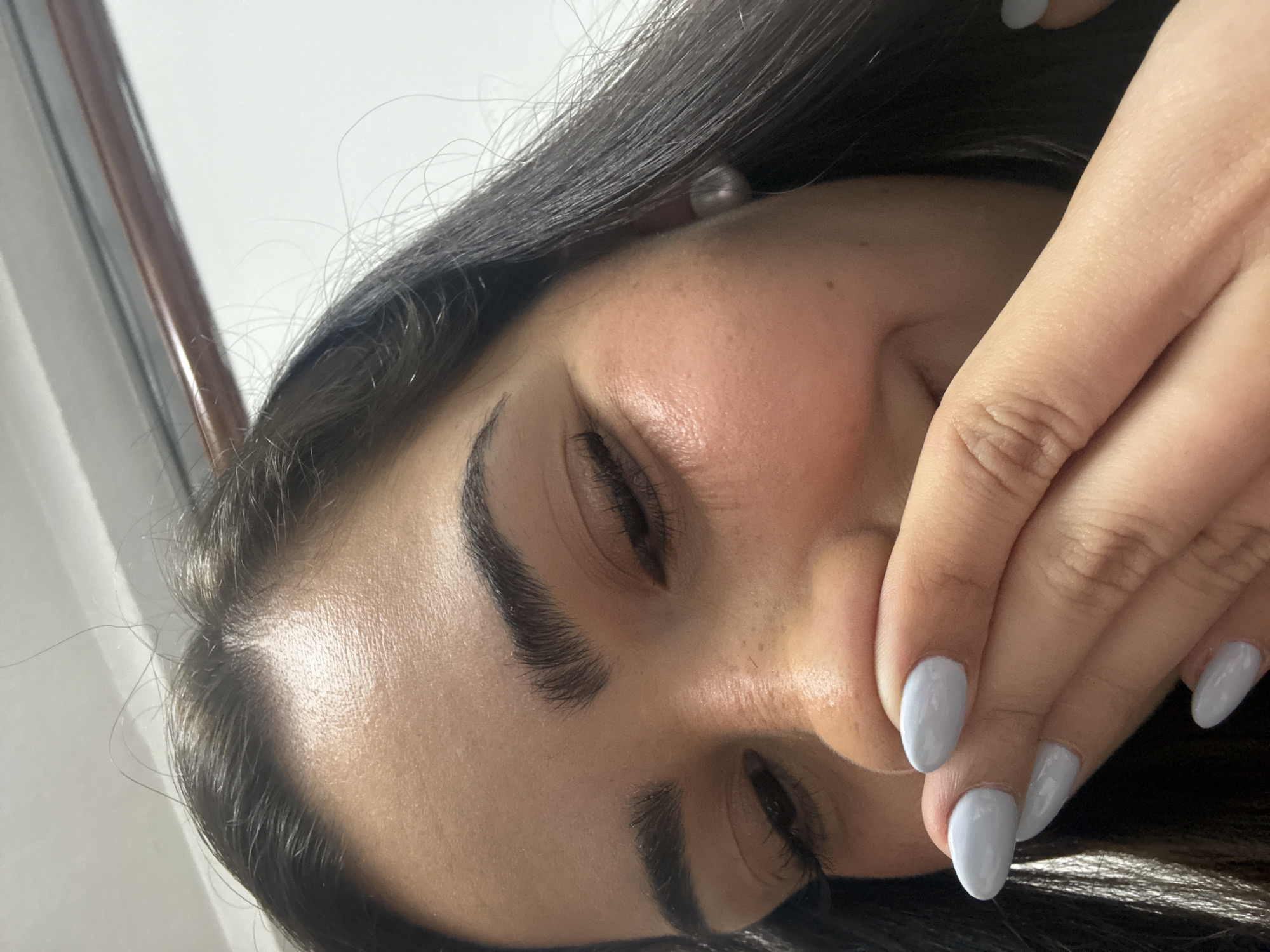 The image shows a close-up view of a person's face, positioned at an angle. The person has dark hair, well-defined eyebrows, and their eyes are partially closed. Their hand, with fingernails painted a light gray color, is held near their mouth, obscuring the lower portion of their face. The skin appears to be fair-toned. The background is out of focus and appears to be a white wall, with a portion of a window frame visible on the left. The location in Puebla, Mexico is not discernable from the details in the photograph.