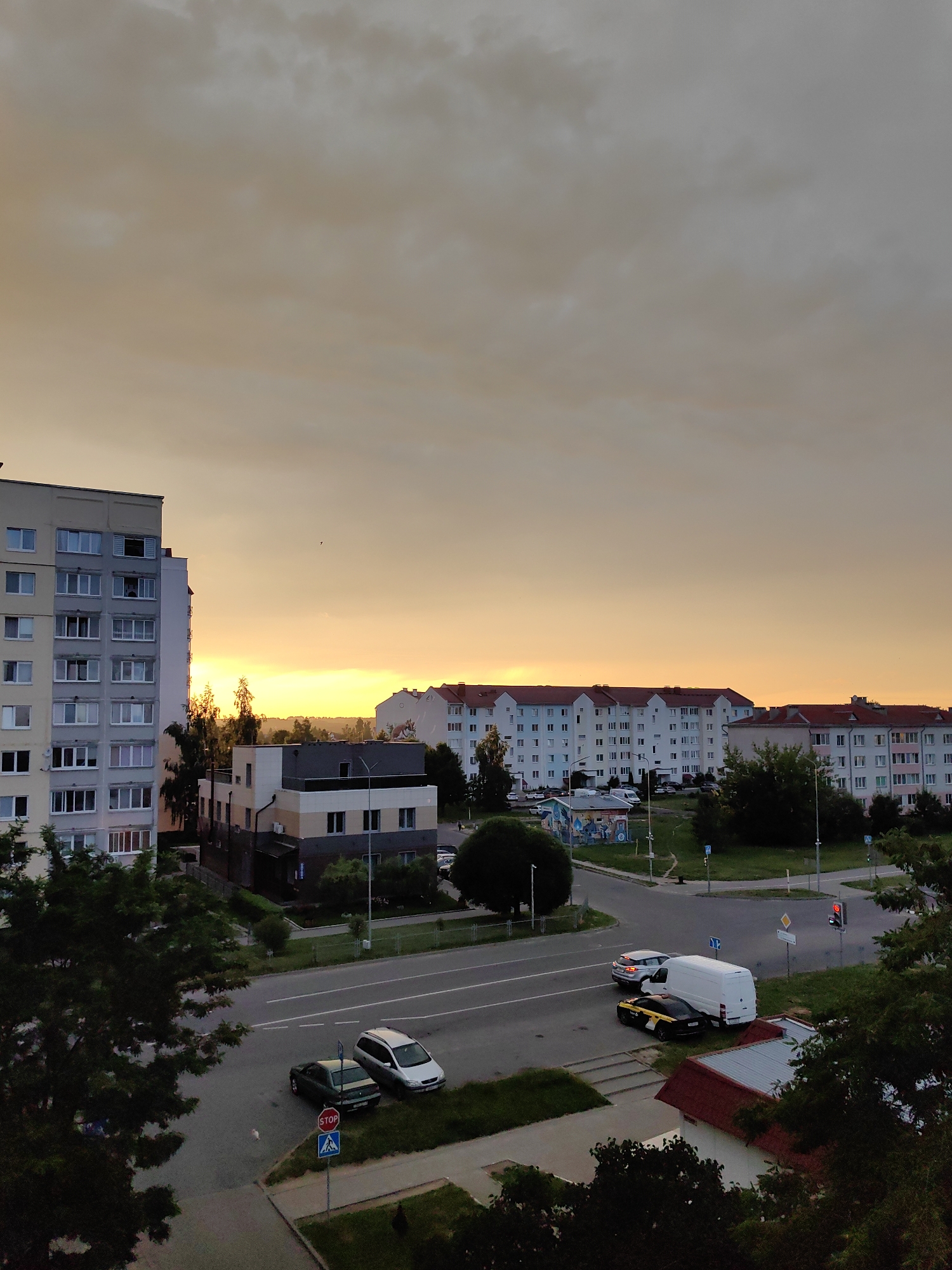A high-angle, wide shot captures a view of a city street in Fanipal, Belarus, during a sunset. The sky is a gradient of orange and yellow, with scattered clouds. In the foreground, a large apartment building is visible on the left, with multiple windows and balconies. Across the street, other apartment buildings with red-tiled roofs are lined up. Several cars, including a white van, a black and yellow car, and a silver SUV, are parked or driving on the road. Trees and green spaces are interspersed throughout the scene. A stop sign is visible at a street intersection. The overall impression is of a residential urban area at dusk.