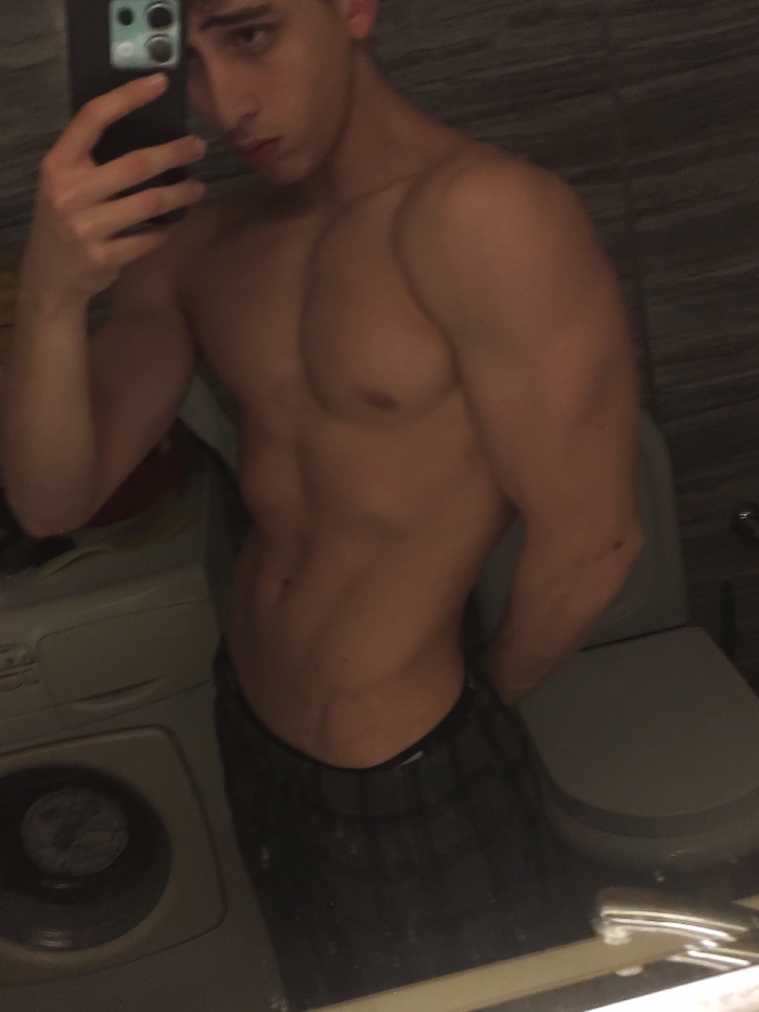 A shirtless young man takes a selfie in a bathroom mirror. He has a muscular physique with defined pectoral and abdominal muscles. He is wearing plaid shorts or boxers. A washing machine is visible to the left, and a toilet is seen behind him to the right. The lighting is dim, creating shadows that accentuate his musculature. He is holding a smartphone up to take the picture.