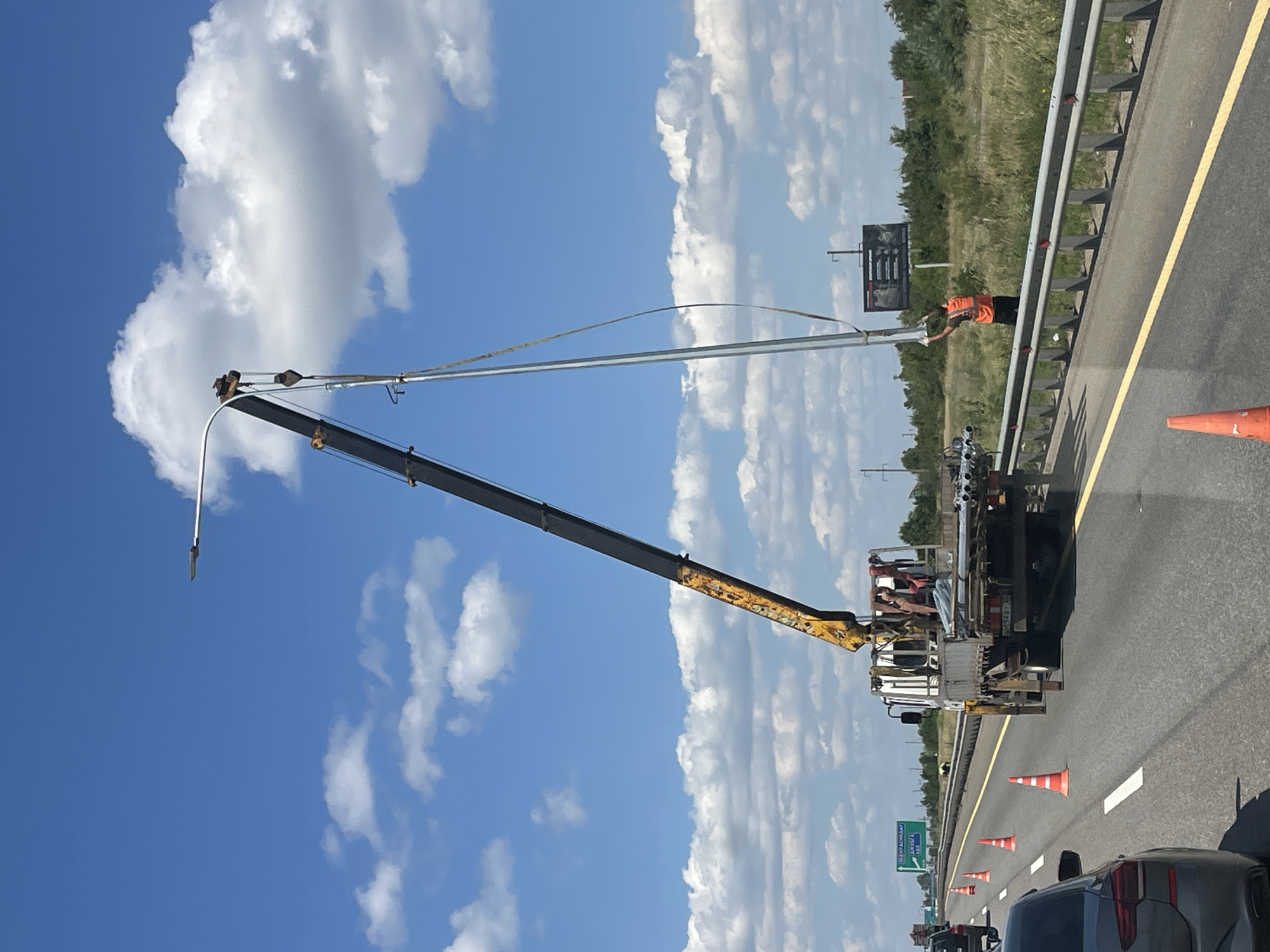 On a highway in Molkino, Russia, a crane truck is installing a new street light pole. The truck is positioned on the side of the road, with its crane extended to lift the pole. A worker in an orange vest is guiding the pole into place, while another person is visible inside the truck's cab. Orange traffic cones are placed around the work area to alert other drivers. In the background, there is a billboard and a tree line. The sky is blue with scattered clouds.