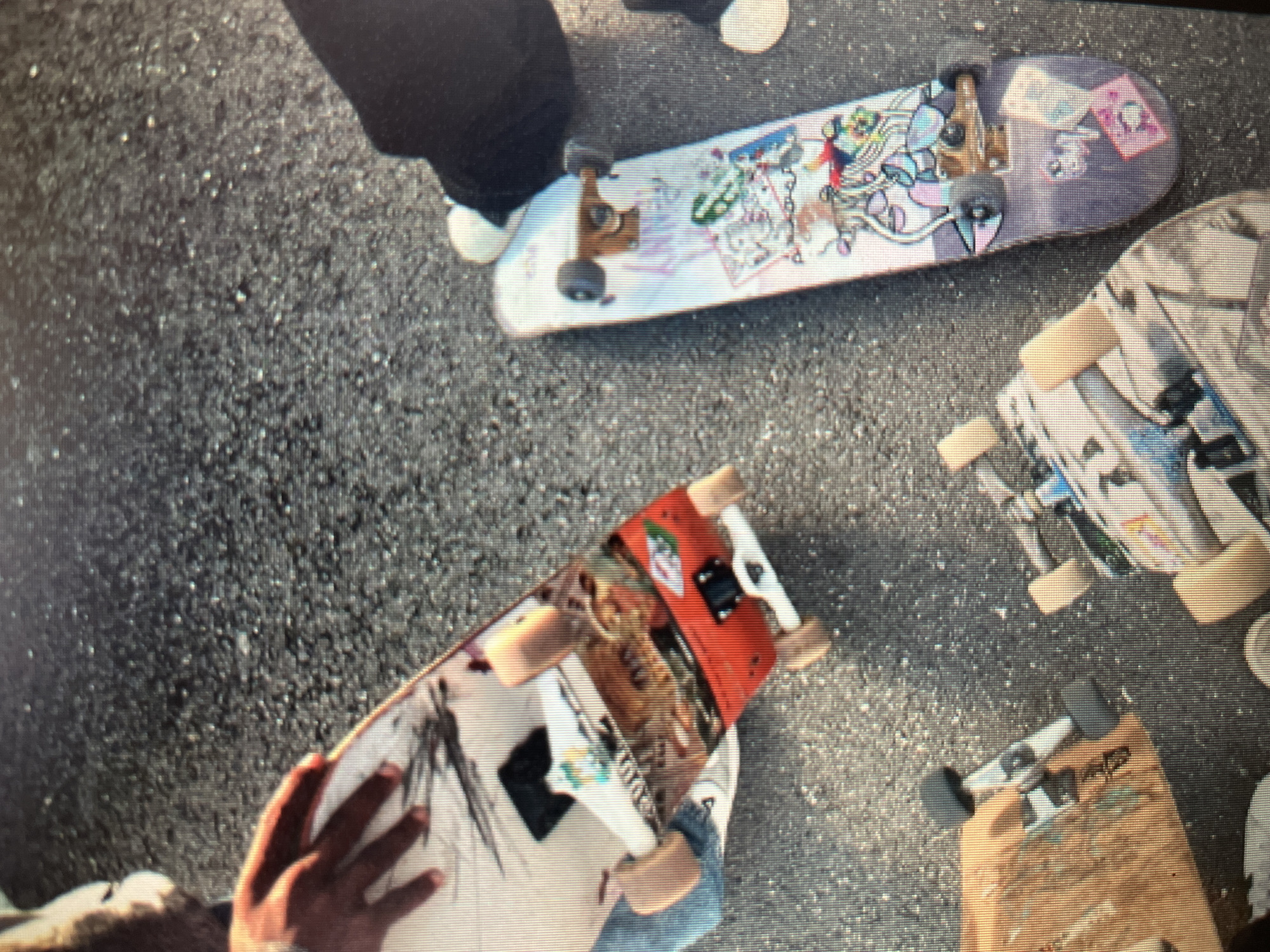 The image captures a ground-level view of several skateboards resting on asphalt. The scene appears to be outdoors, possibly in a public space like a park or street, with the textured grey asphalt suggesting an urban environment. One skateboard in the foreground is predominantly white with graphic illustrations, including what appears to be a black sketch of a bird or insect, and other colorful, fragmented imagery. This board has orange wheels and silver trucks. Next to it, a second skateboard is positioned diagonally, its deck covered in a collage of stickers and graphics, featuring cartoonish figures and text. This board has purple accents on its graphic design and appears to have brown trucks and dark grey wheels. To the left, another skateboard is partially visible, its wooden deck showing a worn, natural finish with some graffiti-like markings. It has light-colored wheels and silver trucks. A fourth skateboard, the closest to the viewer's bottom right, also exhibits a graphic-heavy design with various stickers. The image does not provide any specific landmarks or identifiable features that would place it in Sejahtera, Indonesia. There are partially visible human hands and legs wearing casual clothing, indicating the presence of people, likely skateboarders, who are either about to use the boards or have just finished. The lighting suggests it is daytime.