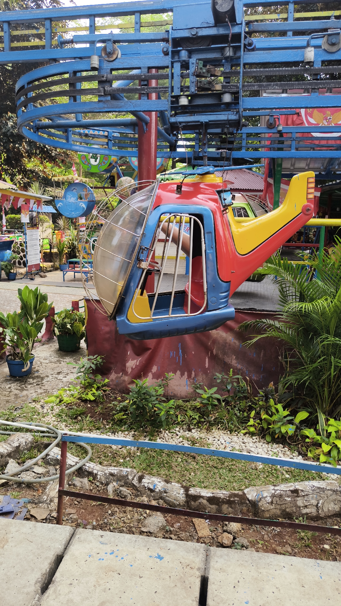 In an amusement park in South Jakarta, Indonesia, a colorful toy helicopter attraction is captured from a slightly low angle. The helicopter, painted red, yellow, and blue, is suspended from a blue metal track, suggesting it's part of a ride. The cockpit of the helicopter is visible, and a person's arm and shoulder can be seen inside, indicating someone is occupying the ride. The background reveals more of the amusement park's infrastructure, including more blue metal structures, trees, and foliage, hinting at a vibrant and playful environment. The ground is paved with concrete and gravel, with some scattered greenery and a low fence in the foreground. The overall scene depicts a typical day at a local amusement park.