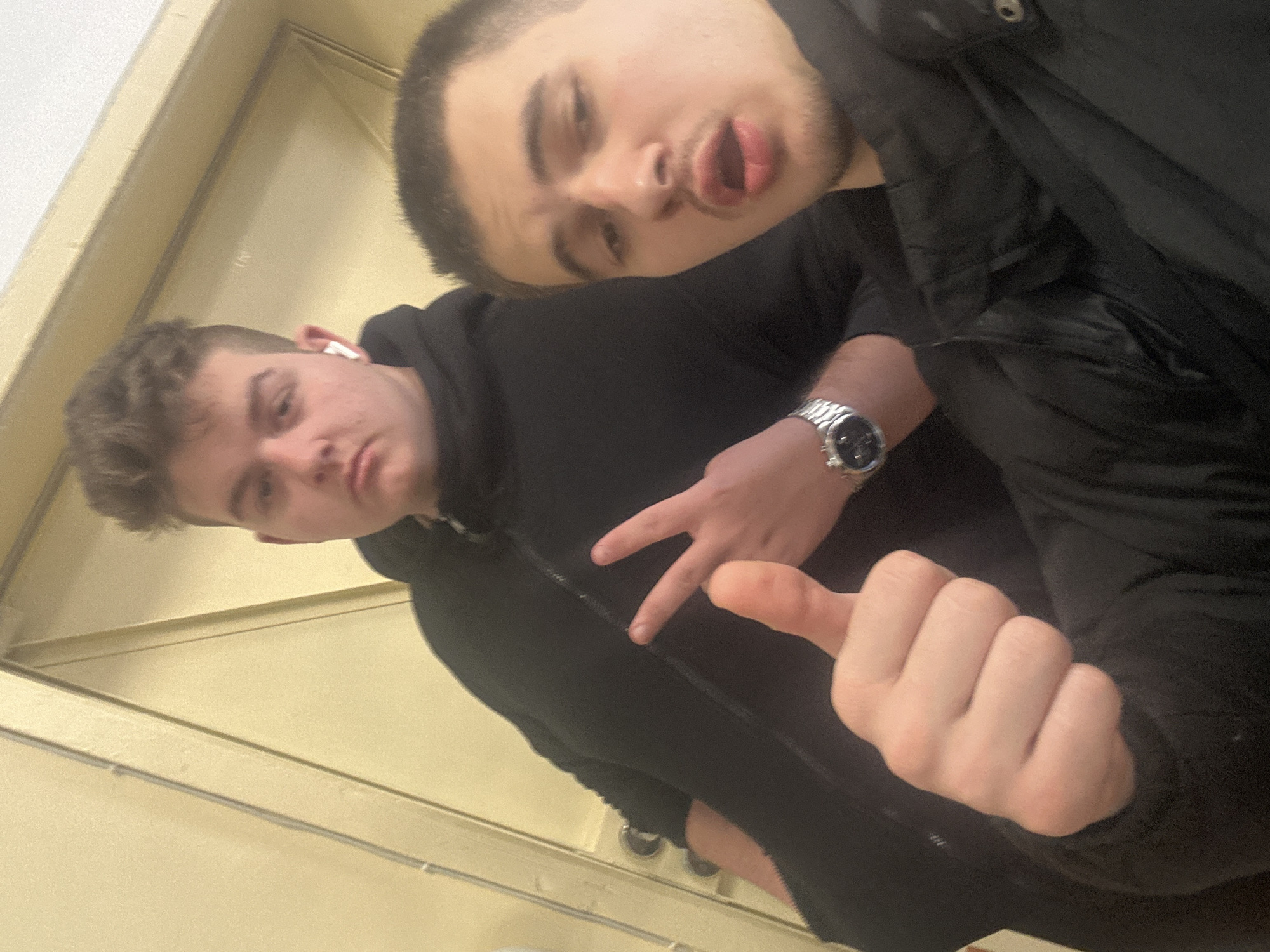 Two young men are posing for a selfie indoors. The man in the foreground is wearing a black hoodie and giving a thumbs-up sign. He has an earpiece in his left ear. The man in the background is making a kissing face. Both men are wearing black clothing. One of the men is wearing a watch. The background appears to be a wall or door with light-colored trim. The specific location in Sofia, Bulgaria is indeterminable with the provided information.