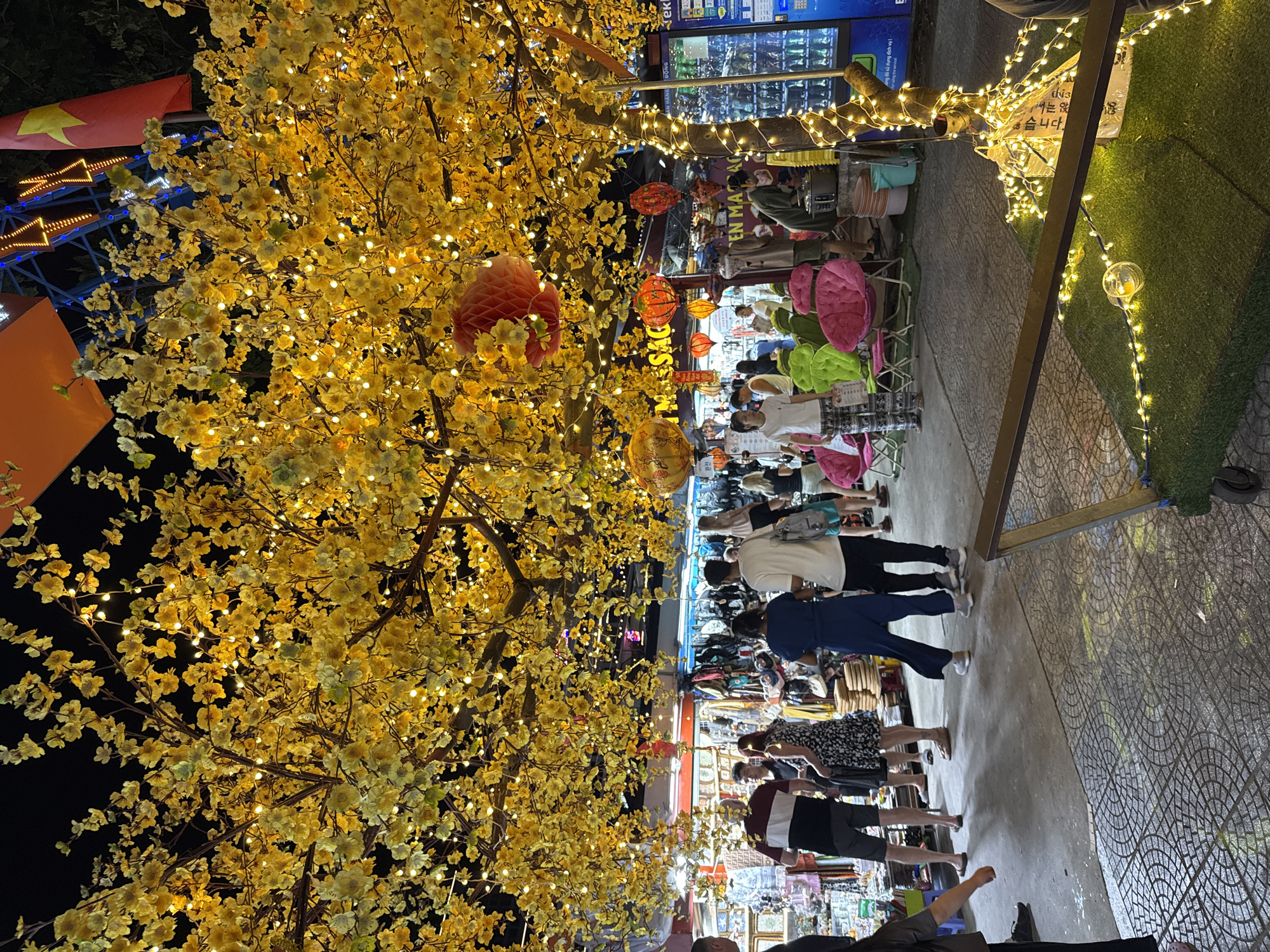 The image captures a lively night scene in Đà Nẵng, Vietnam. The scene is predominantly lit by strings of warm-toned lights adorning trees and stalls, creating a festive atmosphere. Numerous people stroll along a tiled pathway lined with shops and stalls selling various goods. There are red and yellow lanterns hanging adding to the ambiance. A vendor machine with the words "Ekic" is also seen. The image shows the movement of the shoppers as they browse the merchandise. A Vietnamese flag is partially visible, adding to the local flavor.