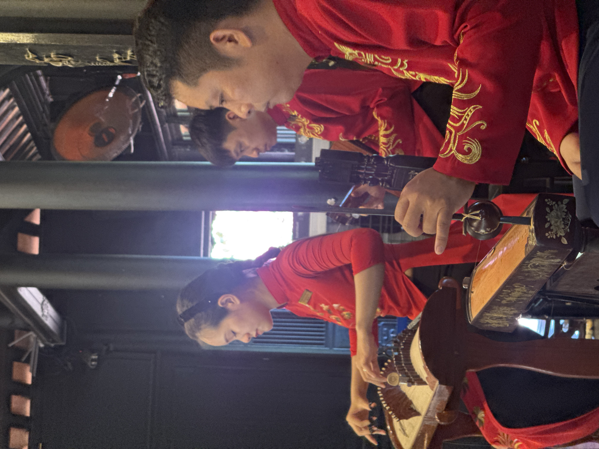 In the city of Bac Nha Trang Ward, Vietnam, three individuals, two men and one woman, are positioned in what appears to be an indoor performance or rehearsal space. The woman, situated on the left, is actively playing a traditional stringed instrument, possibly a zither or guzheng, with both hands. She is wearing a vibrant red garment adorned with floral embroidery and has her hair pulled back with a black headband. To her right, two men, also in matching red attire with gold embroidery, are visible. The man in the middle is partially obscured, while the man on the far right is in profile, his gaze directed away from the camera. The background suggests an architectural setting with dark pillars and a ceiling structure, and a large, orange fan is mounted overhead. The lighting is somewhat dim, with a bright light source to the right, creating a contrast in the scene.
