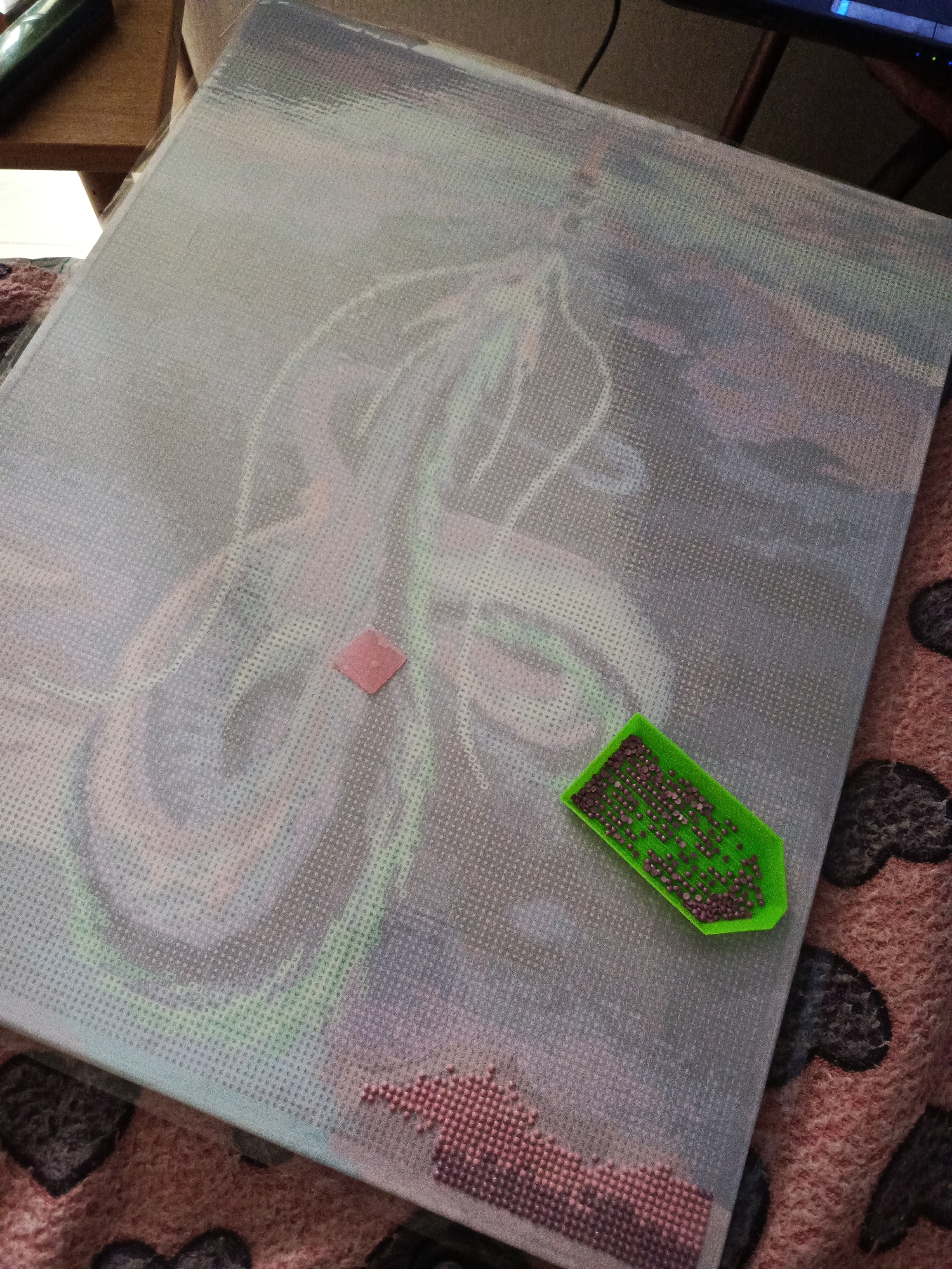 The image displays an overhead view of an ongoing diamond painting project set in an indoor environment, possibly a home in Chaikovsky, Russia. A large, rectangular canvas, covered partially by a translucent protective film, rests on a patterned blanket. The canvas depicts a partially completed artwork, which appears to be a stylized creature, possibly a dragon or serpent, with prominent green outlines and filled sections in various shades of purple, green, and blue. The grid pattern for diamond placement is clearly visible on the unworked areas of the canvas.
In the lower-right section of the canvas, a completed area of densely packed purple-pink diamond drills is visible. A small, vibrant green plastic tray, filled with numerous loose, round, purple-pink diamond drills, is positioned on the canvas. Adjacent to the central area of the artwork, a small, square piece of pink wax, typically used to pick up the drills, is placed directly on the canvas.
The blanket beneath the canvas features a dark, abstract pattern, resembling hearts or clouds, against a reddish-brown background. In the background, to the upper-left, a wooden surface, likely a table or shelf, is partially visible. To the upper-right, a dark electronic screen, possibly a computer monitor or television, is faintly discernible. The lighting is soft and even, typical of an indoor setting during daylight hours. No visible text is present in the image.