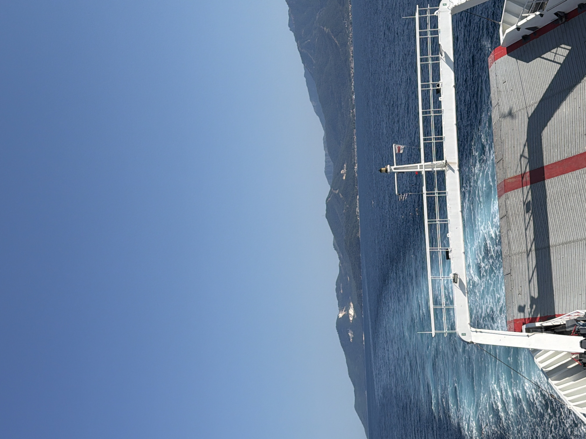 This is a vertical, medium-shot photograph taken from the deck of a ferry, looking out towards a coastline with mountains. The sky is a clear, bright blue, indicating a sunny day. The sea is a deep blue, with white wakes trailing behind the ferry.
The ferry's structure is visible on the right side of the image, with grey metal siding and red stripes. A white metal railing runs along the side of the ferry, extending into the water. Two flags are visible on a pole attached to the ferry: one is a white flag with a red cross, and the other appears to be the Greek flag. The shoreline, characterized by lush green mountains and some scattered buildings, is visible in the distance on the left.
The overall scene suggests a leisurely journey on a ferry under clear skies. The lighting indicates it is daytime. The location is Keramoti, Greece, which aligns with the mountainous coastline and the presence of the Greek flag. There is no visible text in the image.