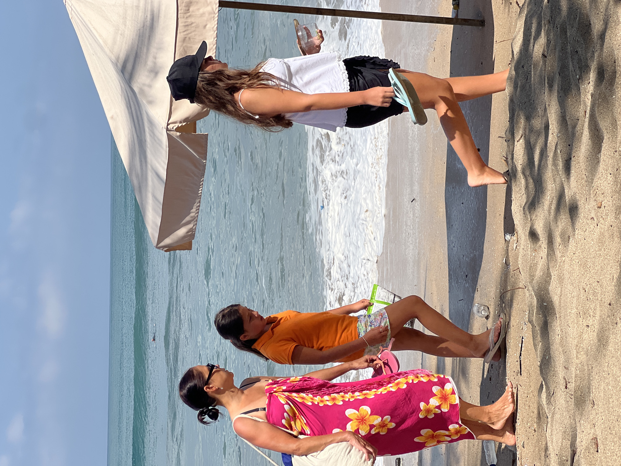 In Kuta, Indonesia, on a sunny day at the beach, three individuals are walking along the shoreline. A woman in a bright pink sarong with yellow and white floral patterns is on the left, her hair pulled back in a bun, wearing sunglasses. Beside her, a young girl in an orange polo shirt and patterned shorts walks with her gaze lowered. To the right, a third person, possibly a teenage girl, is wearing a white tank top, black shorts, and a black baseball cap. She is carrying a clear container and a light blue flip-flop in her hand. They are all walking away from the viewer, towards the right side of the frame. The ocean is visible in the background with gentle waves. A large, light-colored umbrella provides shade on the right side of the image. The beach sand is light brown, with some debris scattered across it.