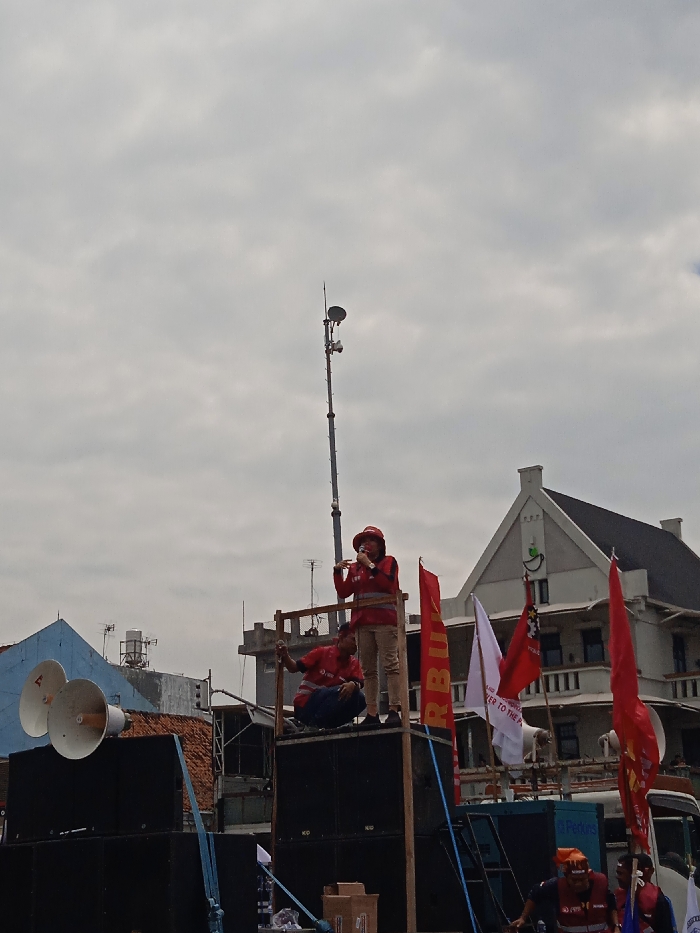 The image was taken in Yogyakarta, Indonesia, at the coordinates -7.783, 110.367. Two individuals are standing on a stage made of wooden planks and large speaker boxes. One is speaking into a microphone. Both individuals are wearing red shirts and reflective vests. Behind them are red and white flags. The stage is in front of a building with a light gray facade, a green roof, and white window frames. On the left side of the image, there is a large speaker system with two blue speaker horns. In the background, a tall metal pole with a satellite dish and other equipment at the top can be seen. There are also various wires and cables visible in the image. The sky is overcast and gray.