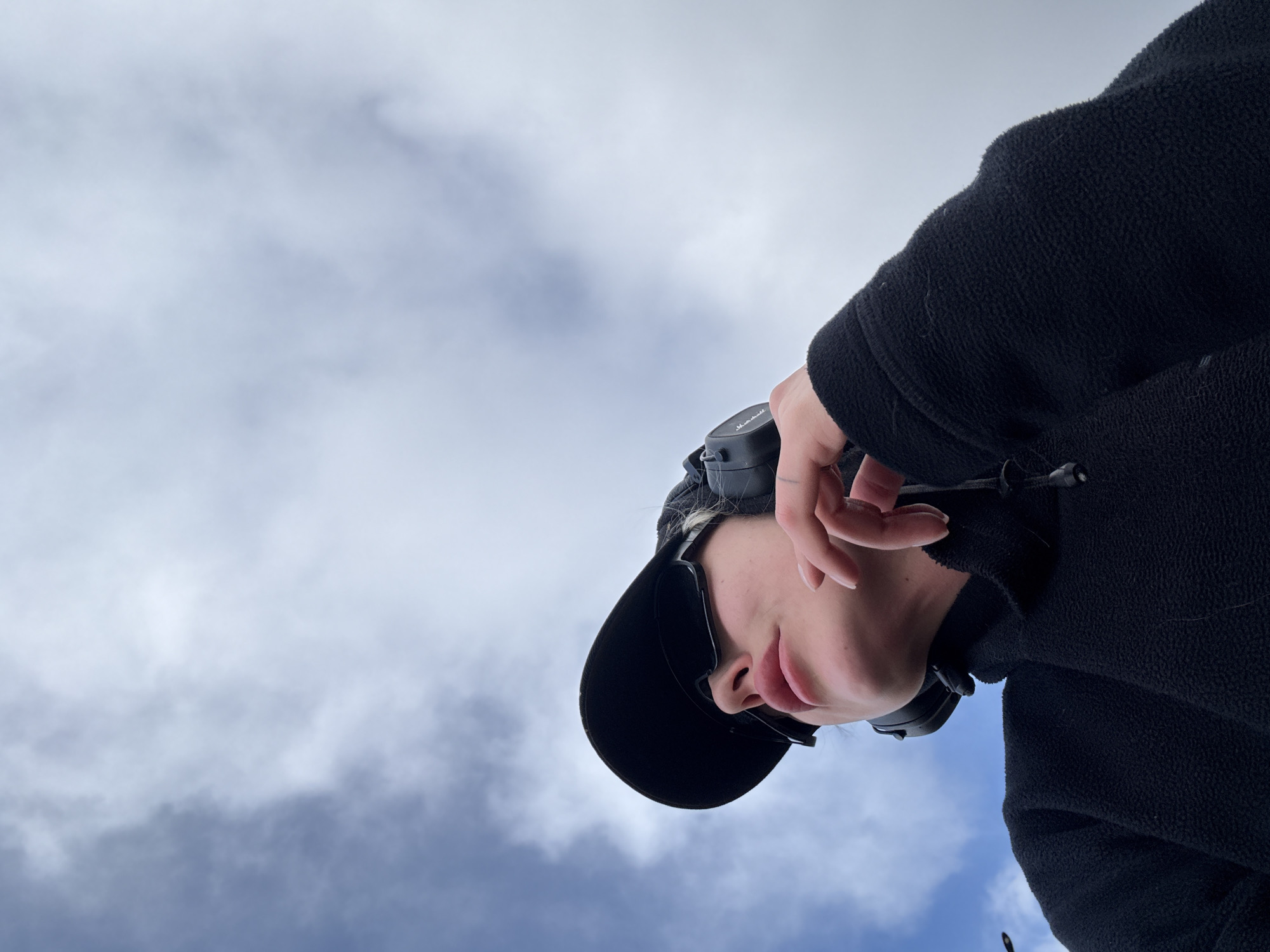An outdoor, low-angle shot captures a person against a cloudy sky. The individual, positioned in the bottom right portion of the frame, is seen from a worm's-eye view, looking slightly downwards or to the side. They are wearing a black fleece jacket, a black baseball cap, and black over-ear headphones, which bear the "Marshall" brand logo on the ear cup. Their face is partially obscured by the cap, but their chin and mouth are visible, with lips slightly parted in a neutral or relaxed expression. One hand is raised towards their face, with fingers gently curled. The background consists entirely of a sky filled with a mix of white and grey clouds, suggesting an overcast or partly cloudy daytime. This scene is set in Krasnoyarsk, Russia.