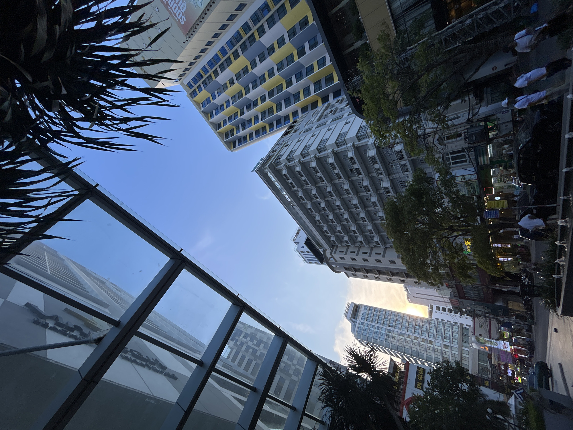 A low-angle shot in Nha Trang, Vietnam, captures the architectural diversity of the urban landscape. Looking upwards, the scene is framed by a geometric glass structure on the left and the spiky leaves of a palm tree on the upper left. Skyscrapers of varying architectural styles rise into the blue sky, with one showing a modern design with yellow and grey accents. The right side of the image displays a street scene with buildings, trees, and people walking. The sun is partially visible, casting a warm light on the cityscape.
