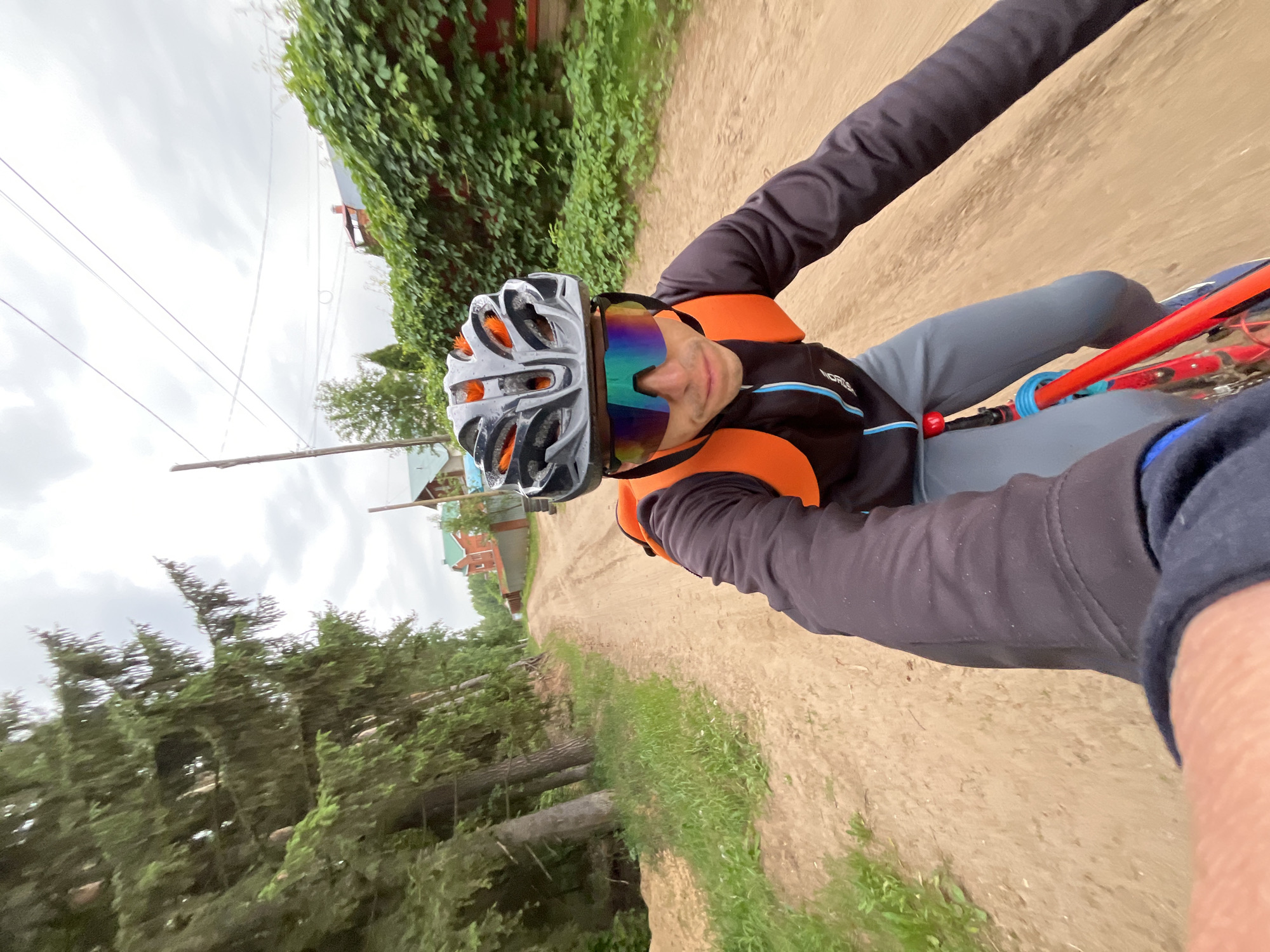 The image shows a person cycling on a dirt road in Chaikovsky, Russia. The individual, likely male, is positioned in the center, facing the camera for what appears to be a selfie. They are wearing a grey and orange cycling helmet and large, multi-colored reflective sports sunglasses. Their attire includes a black long-sleeved athletic top, a black and bright orange sleeveless vest or jacket, and grey pants. A logo that reads "NORTH" with some additional small text or design is visible on the chest of their top.
The person is seated on a bicycle, with parts of a red bike frame and some blue and red accessories visible near their leg. The road is unpaved, made of light brown dirt, with patches of green grass along its edges.
On either side of the road, there is dense green vegetation. To the left, several conifer-like trees are visible. To the right, green vines climb a structure or wall, and beyond them, residential buildings with reddish-brown roofs can be seen. Power lines and utility poles are present against the sky. The sky is overcast, indicating a cloudy daytime setting, possibly late morning or afternoon, without direct sunlight. The overall impression is one of an active outdoor pursuit in a rural or semi-rural environment.