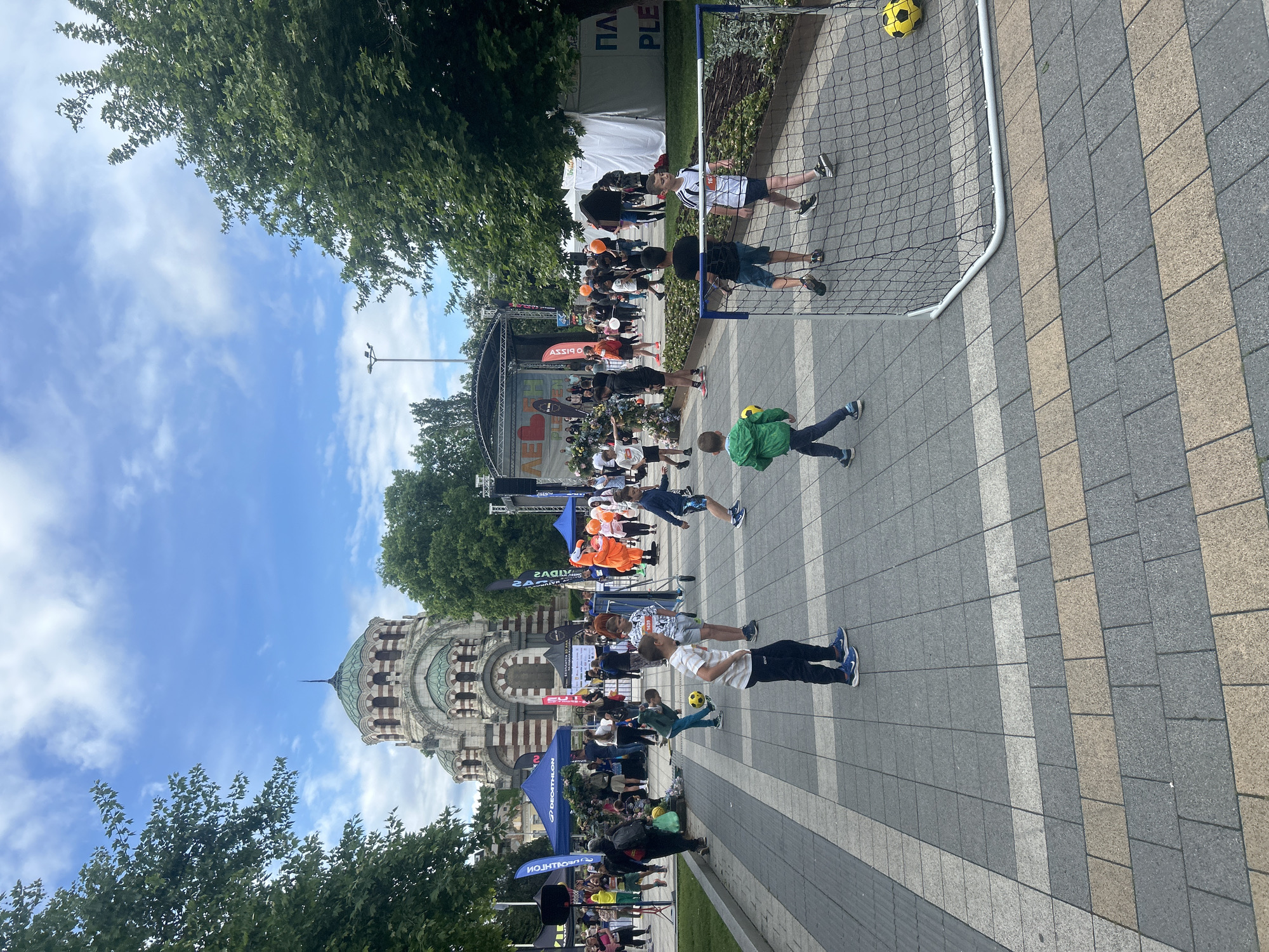 The image shows a scene in Pleven, Bulgaria, with children playing football in front of the Saint George the Victorious Chapel Mausoleum. The chapel is visible in the background, with its distinct architectural style. Several children are actively engaged in a game of football using a small portable goalpost. Other people are standing around or passing through the scene, and there appears to be a small outdoor event or gathering taking place. The environment consists of paved ground, trees, and street furniture.