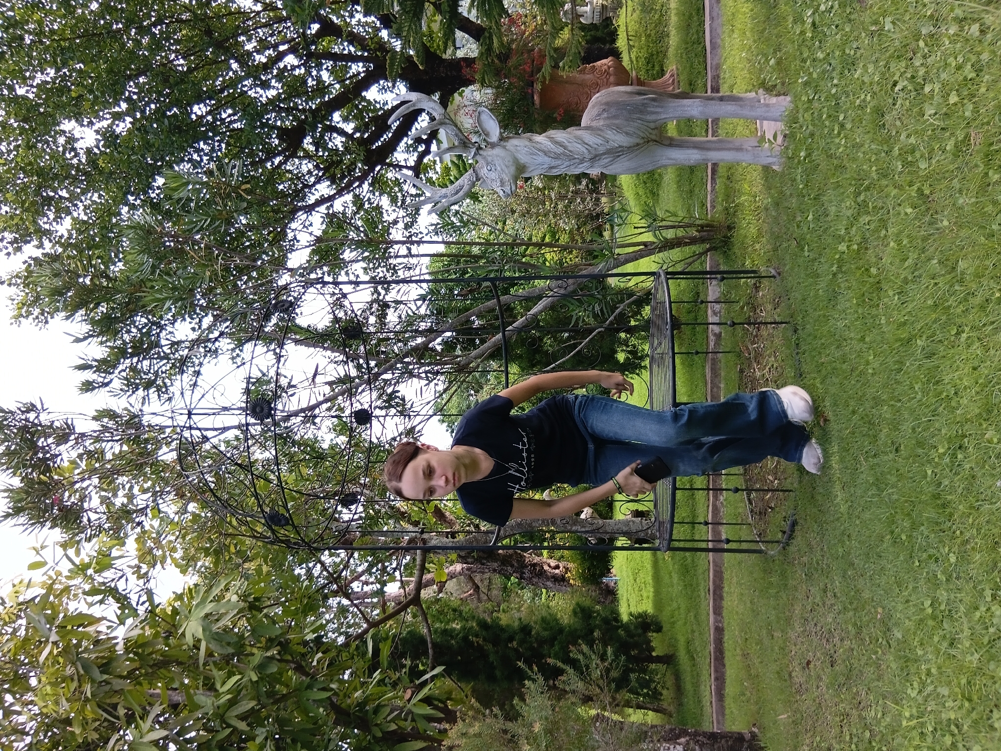 In the city of Ban Tong, Thailand, a young Caucasian female is captured in a photograph outdoors within a garden setting. She is wearing a dark blue t-shirt with white text and blue jeans, with white sneakers on her feet. Her hair is pulled back from her face. She is holding a dark-colored object in her right hand and is leaning forward. To her right, a stone statue of a deer stands prominently. The garden is lush with greenery, featuring trees, bushes, and a well-maintained lawn. A decorative metal structure, possibly a trellis or arbor, is positioned behind the woman. The overall environment suggests a peaceful, outdoor recreational space within Ban Tong.