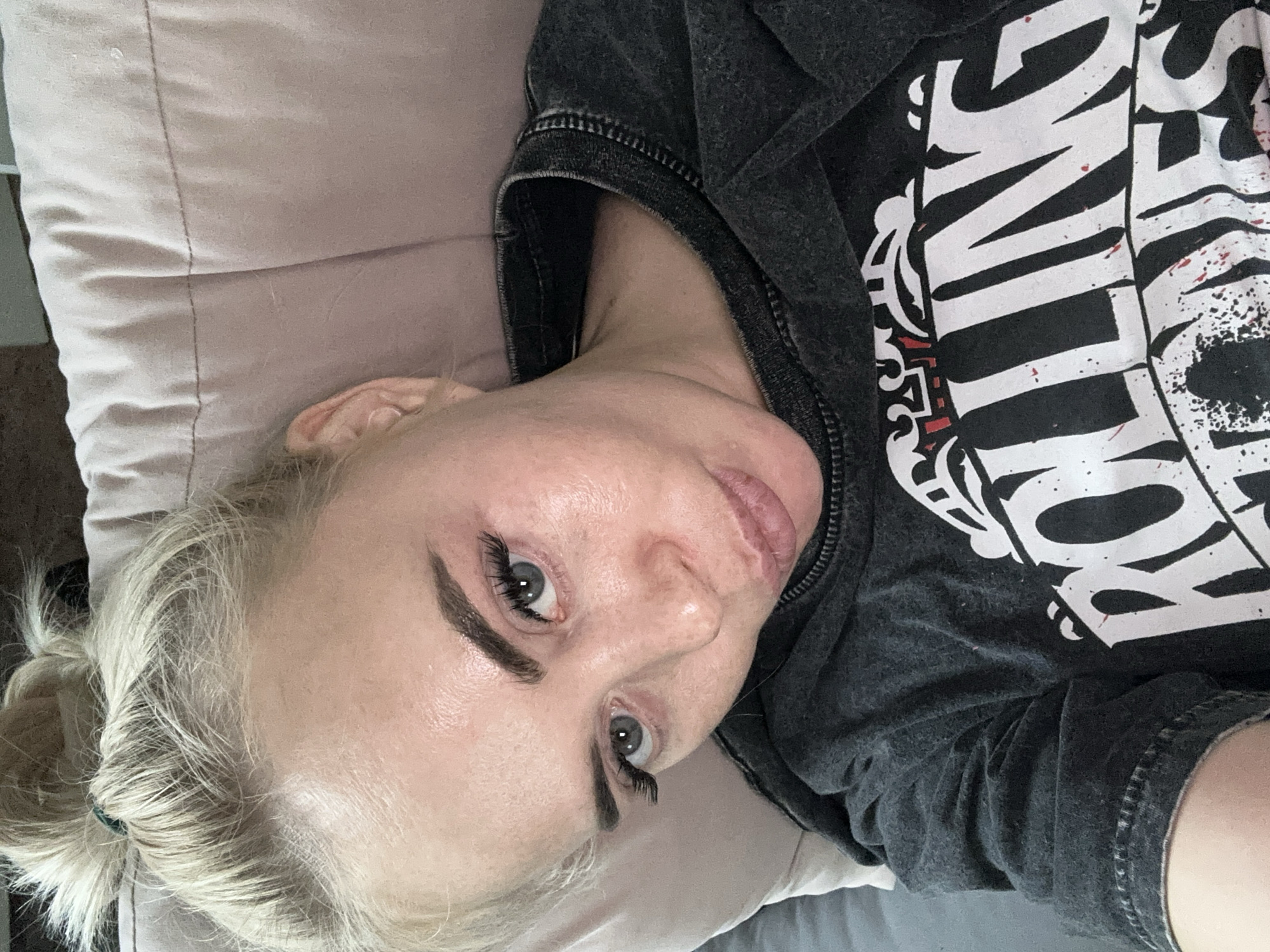 A woman with blonde hair pulled back in a ponytail lies on a bed with a light pink pillow. She is wearing a dark gray t-shirt with a white design. The woman is looking up at the camera. No specific details indicate this is in the city of Высокий, Russia.