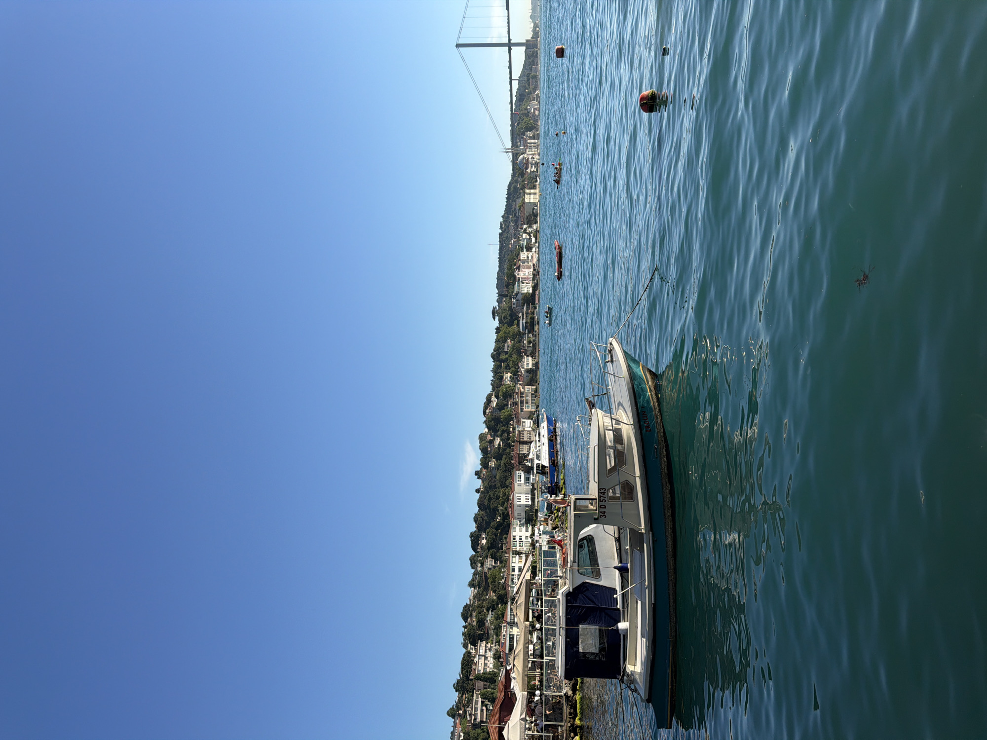 An aerial, eye-level, full shot captures a clear, blue sky over a tranquil bay in Üsküdar, Turkey. In the foreground, a white and blue fishing boat, identified by the license plate "34 D 5743", is anchored to the left, its hull painted a deep teal. The boat is connected to a red and orange buoy further out in the water.
The bay is dotted with other small boats, some carrying passengers, and a few more buoys. In the mid-ground, a bustling waterfront features white umbrellas shading tables where patrons dine, as well as several restaurants. Behind the waterfront, a picturesque hillside is densely populated with white and pastel-colored houses, interspersed with lush green trees.
Dominating the background is the magnificent Bosphorus Bridge, a suspension bridge stretching across the water, its tall pylons clearly visible against the horizon. The bridge connects two sides of the city, a testament to Istanbul's unique geography straddling Europe and Asia. The water is a vibrant turquoise, with gentle ripples suggesting a calm, sunny day. The overall scene evokes a sense of peaceful coastal living, with a backdrop of iconic Turkish landmarks.