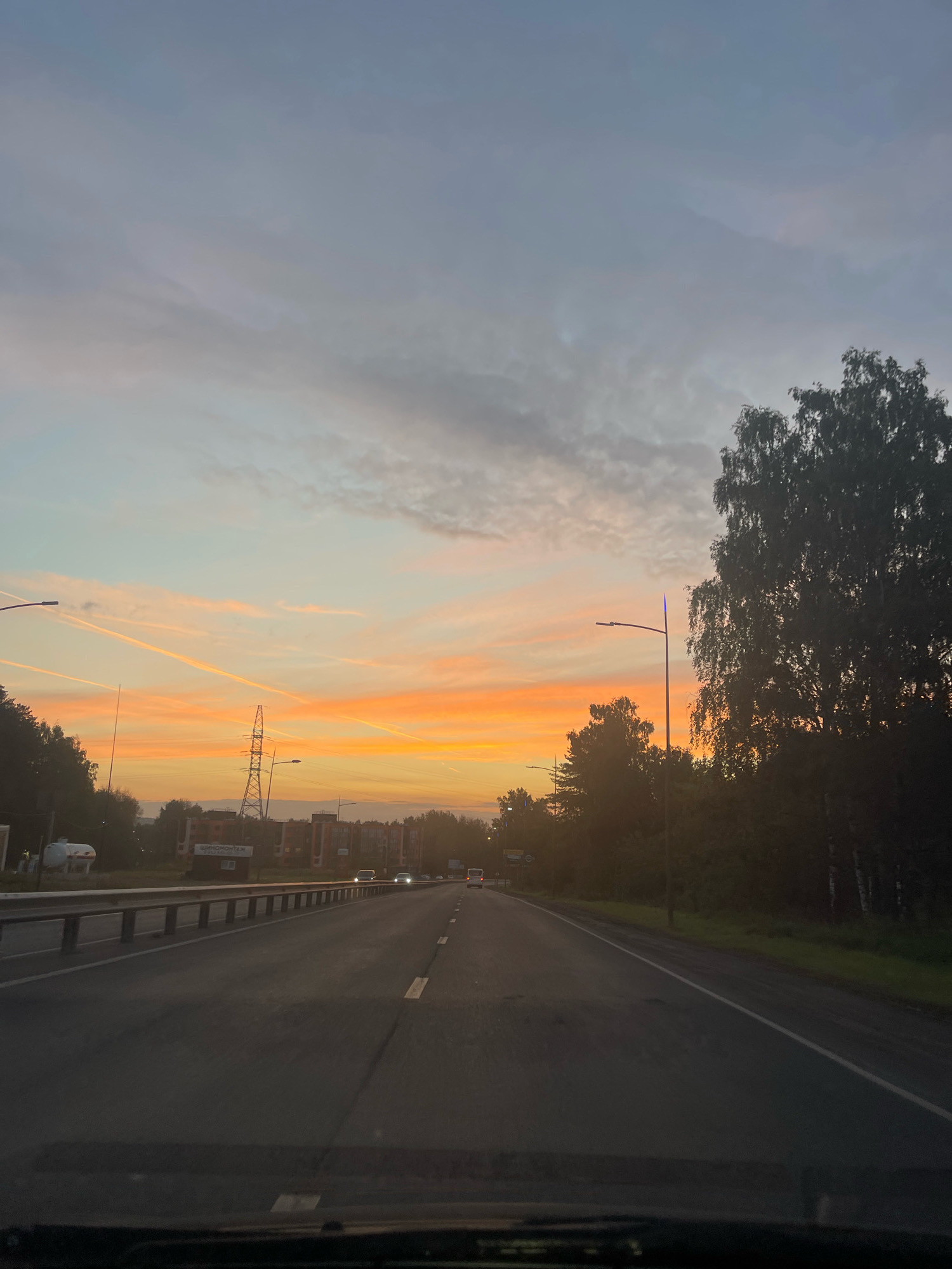 The image depicts a road scene at sunset, likely in the city of Oktyabrsky, Russia, given the Cyrillic text on a sign that reads "ШИНОМОНТАЖ," meaning "tire fitting" or "tire service." The viewpoint is from inside a vehicle, looking forward. A paved, multi-lane road stretches into the distance, with white dashed lines marking the lanes. On the left side of the road, a metal guardrail runs parallel, with trees and some low-rise buildings visible beyond it. A large pylon is prominent in the middle ground, with a sign on a structure beneath it. On the right side of the road, dense trees form a dark line, with a single streetlight visible. The sky above is a gradient of colors, transitioning from a pale blue at the top to vibrant orange and yellow hues near the horizon, indicating the setting sun. Several contrails are visible in the sky, adding a linear element to the scene. A few vehicles are present on the road in the distance. The overall atmosphere is serene and captures the twilight of day.