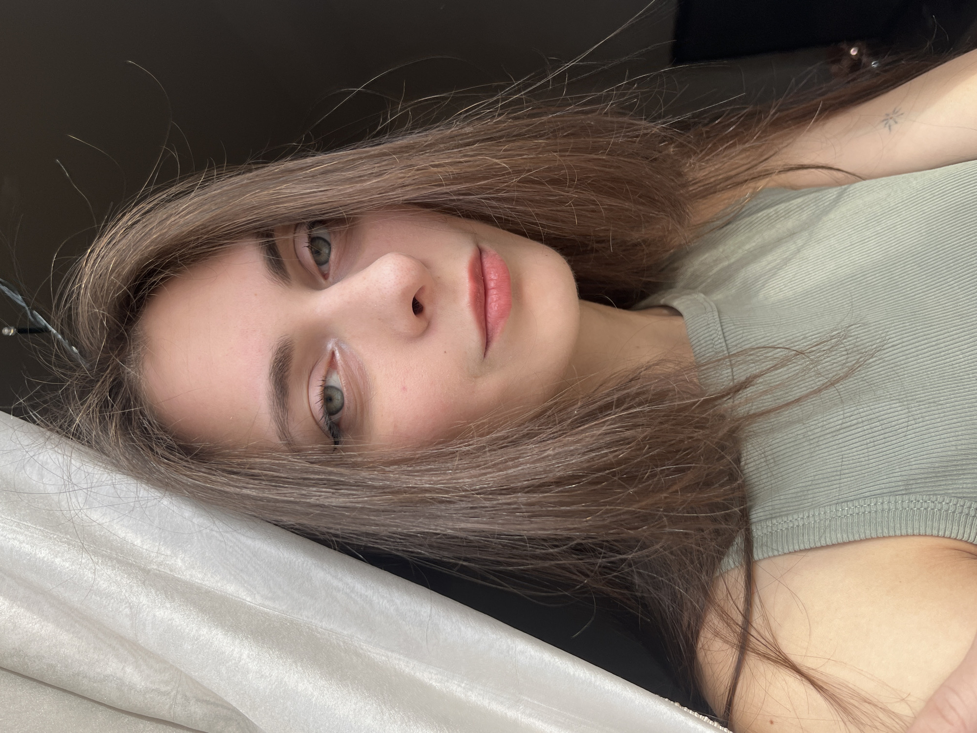 A close-up portrait shows a light-skinned woman with long, brown hair. She is lying down on a white, silk-like fabric, possibly a pillow or bedding. She wears a light green, ribbed shirt. A small tattoo is visible on her upper arm. The background is dark, suggesting an indoor setting. The location is not discernible from the visual information in the image.