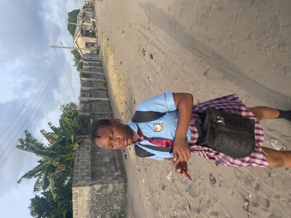 A young female walks on a sandy dirt road in Mosafejaw, Nigeria, under a partly cloudy sky. The subject, appearing to be a student, is centrally framed, facing slightly right, with her eyes closed. She has dark skin and braided hair. She wears a light blue short-sleeved school uniform shirt with red trim and a visible crest on the left chest, a dark red tie, and a red and white plaid skirt. A black backpack is worn on her shoulders, and a small black woven shoulder bag hangs from her left shoulder. She holds a dark red rectangular object, likely a smartphone, with her hands clasped at waist level. To the left, a grey concrete block wall is partially obscured by green vegetation, including banana trees. In the background on the right, a series of single-story, light-colored buildings with corrugated roofs line the road, alongside utility poles and overhead lines. A distant, indistinct human figure is present near these structures. The road surface exhibits tire tracks, footprints, and scattered debris. Long shadows extend towards the right side of the frame.