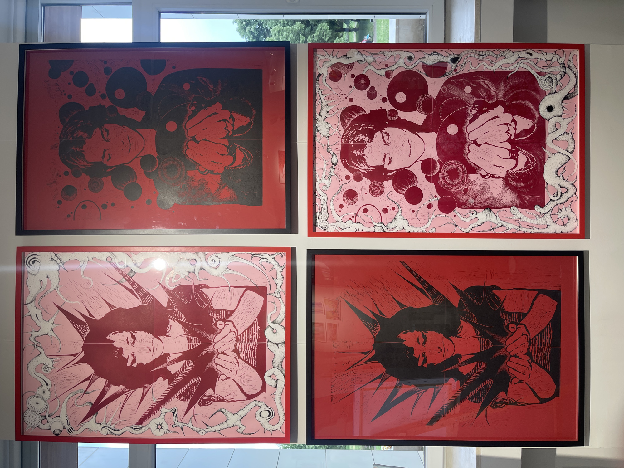 Four framed pieces of art are displayed on a white wall. The art pieces are arranged in a 2x2 grid. The top left and bottom right pieces have black details over a red background and are framed in black. The top right and bottom left pieces are pink and red with white details, framed in red. All pieces depict a person's head and hands, repeated with various shapes and designs around them. Light reflects off the glass of the frames. A window is visible above the art pieces. The city and location are not evident in the image.